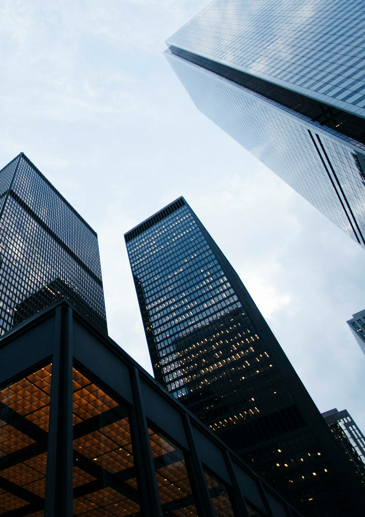 Looking up at tall modern skyscrapers in a city, with reflections on glass facades and a cloudy sky above.