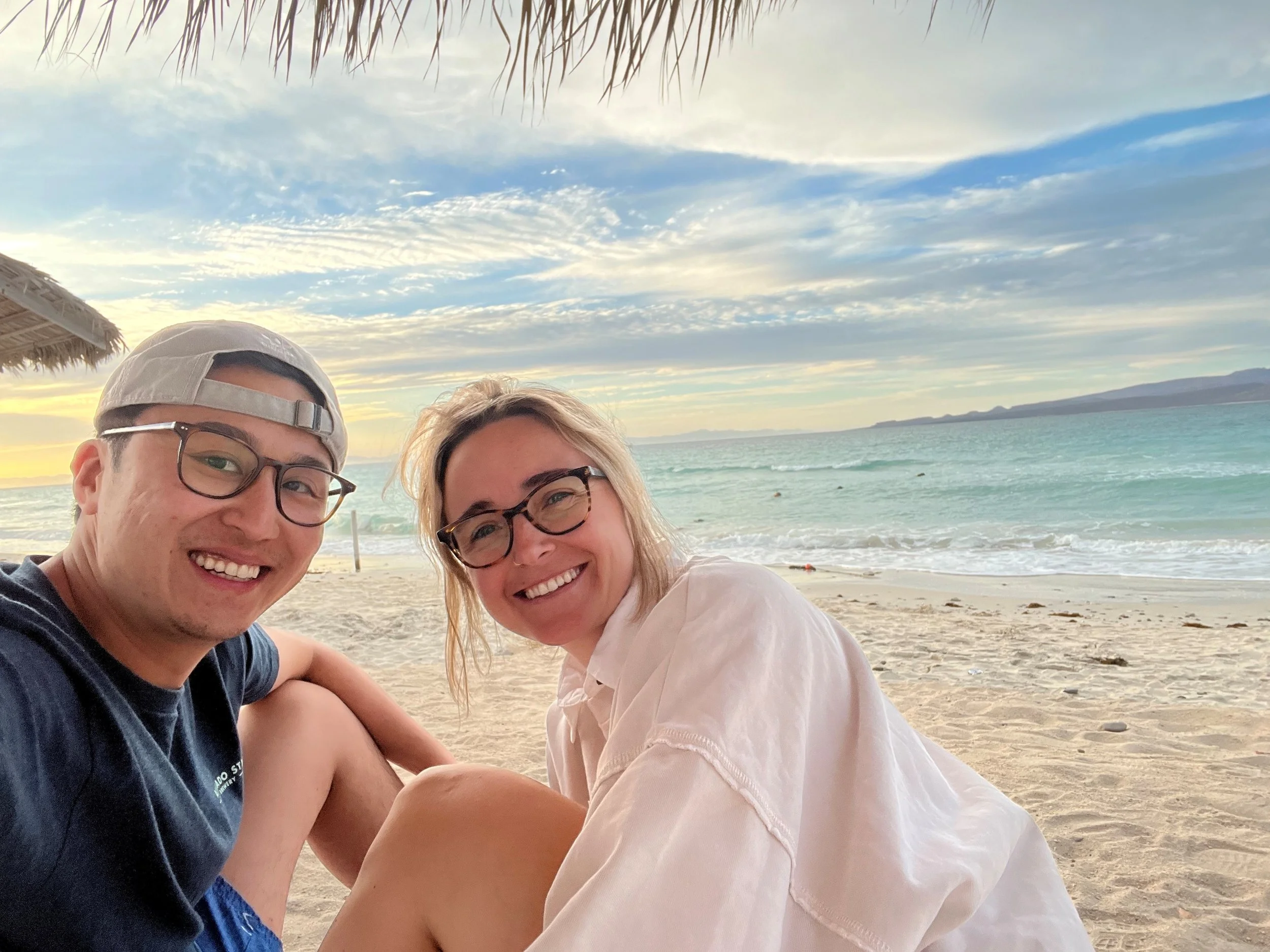A happy couple smiling while sitting on the beach at sunset, with the ocean and sky in the background.