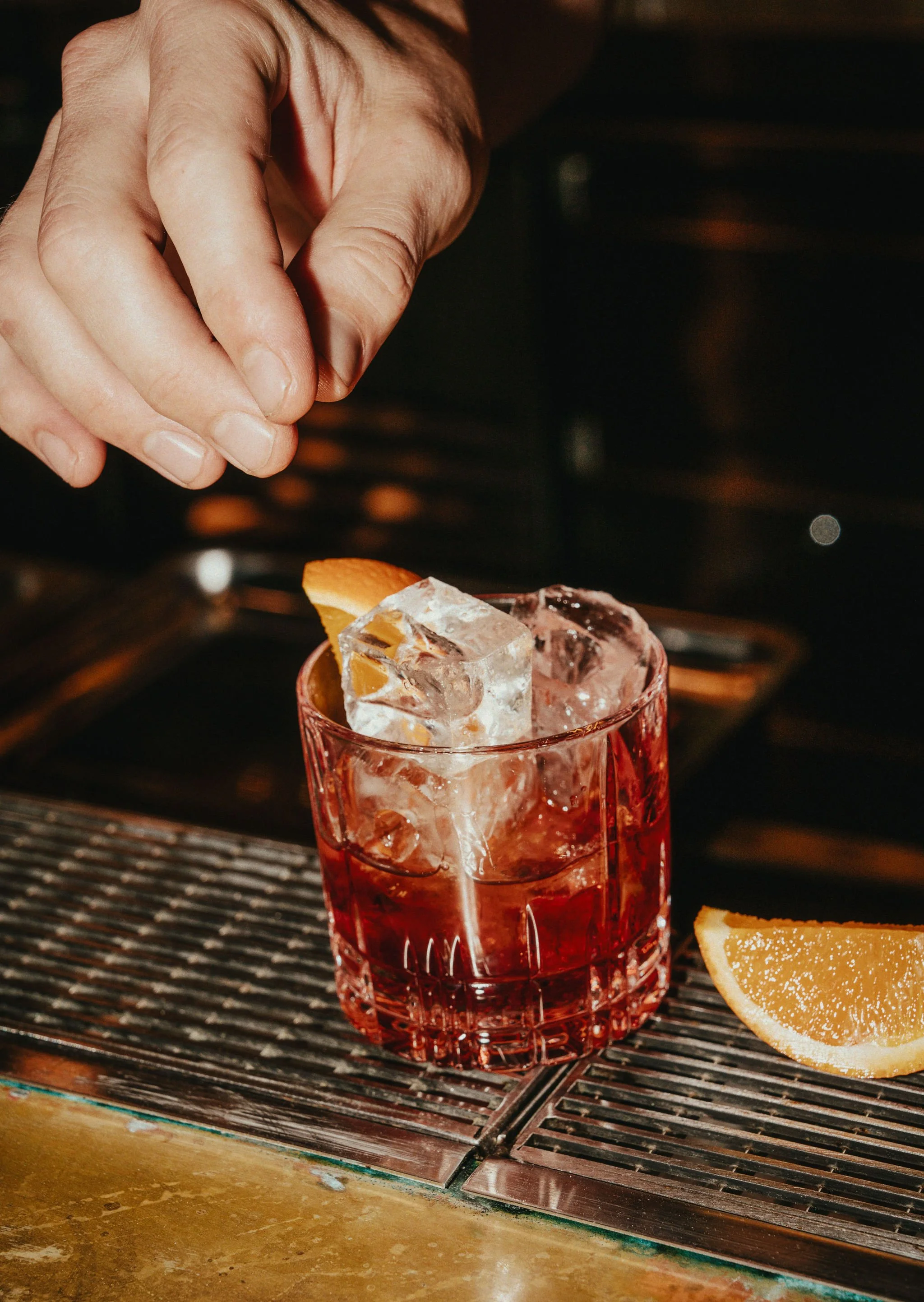A hand garnishing a cocktail with a slice of orange in a red glass with large ice cubes, placed on a bar counter beside another orange slice.