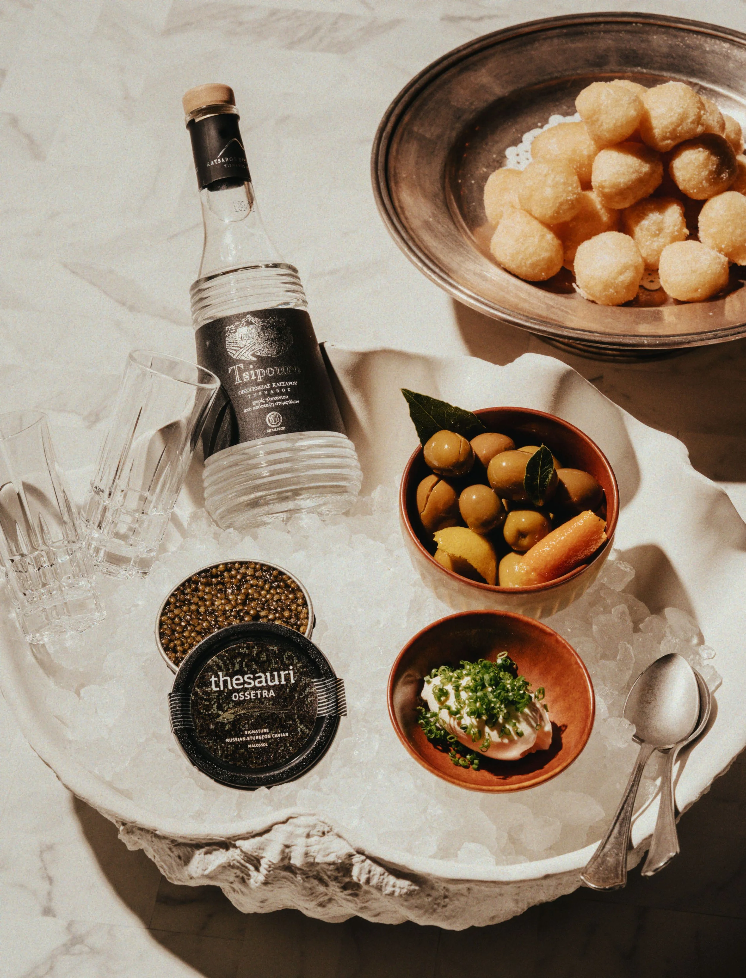 A marble slab displaying a bottle of Tsiropoulos ouzo, two empty glasses, a bowl of caviar, a bowl of olives with lemon and herbs, a bowl of cheese puffs, and a spoon. The setup is on ice with a textured white background.