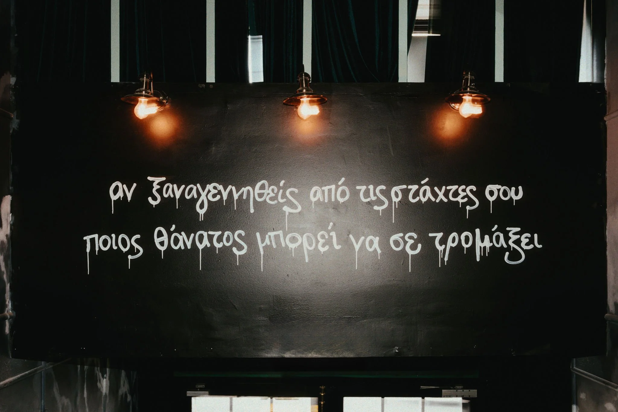 Black sign with Greek writing illuminated by three hanging lights