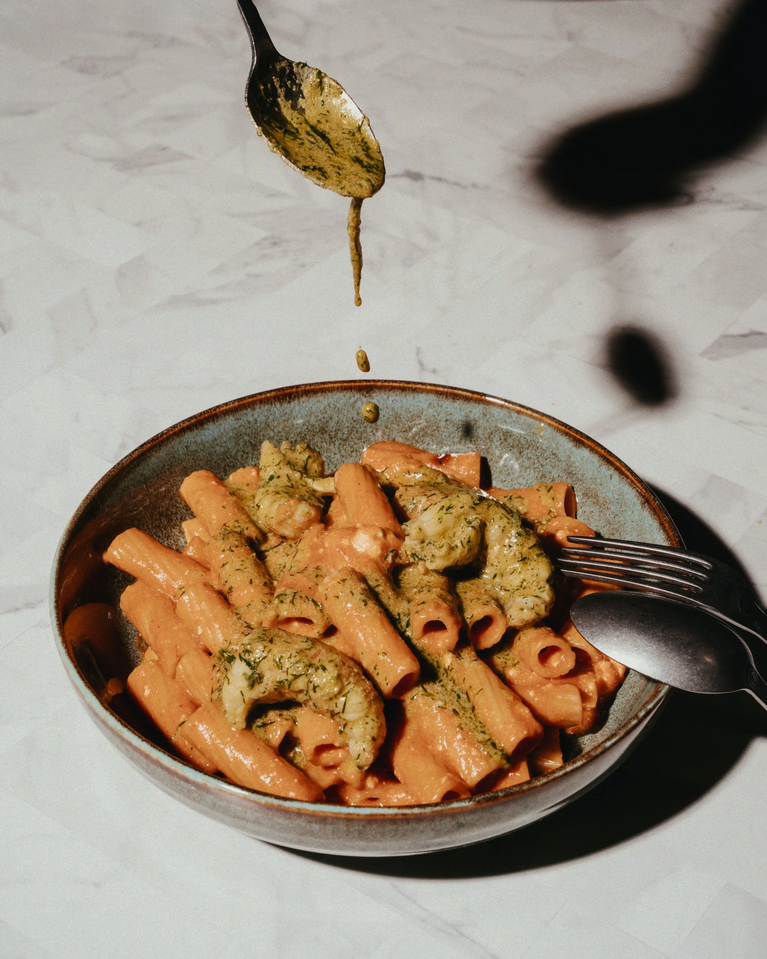 A bowl of cooked pasta with shrimp and green sauce, with a fork and spoon resting on the edge, and sauce being poured over the dish.