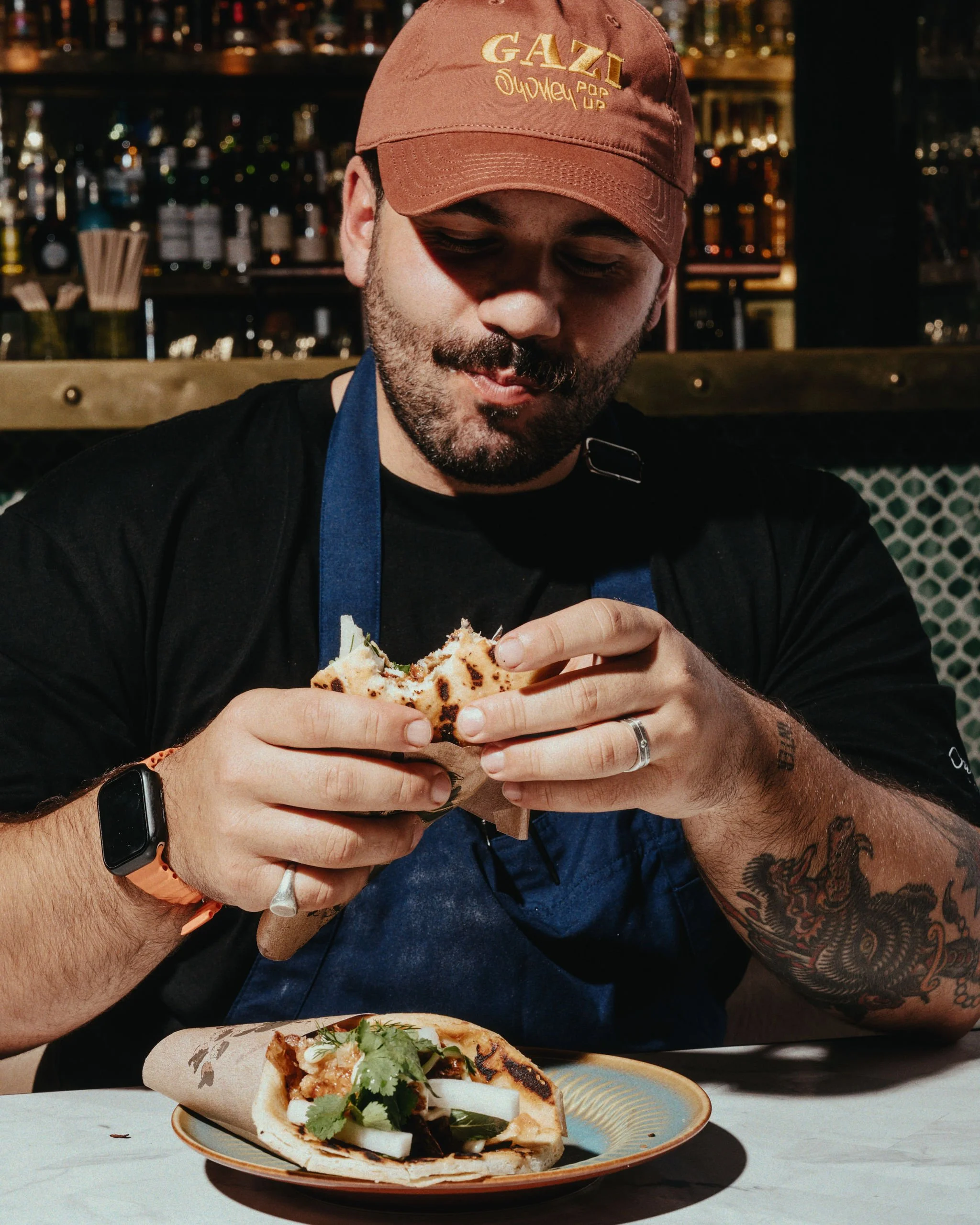 A man with tattoos on his arm, wearing a brown cap and a black shirt, is sitting at a table, holding a slice of pizza and smiling. There is a plate with another type of pizza topped with cilantro, onions, and sauce in front of him, with a background