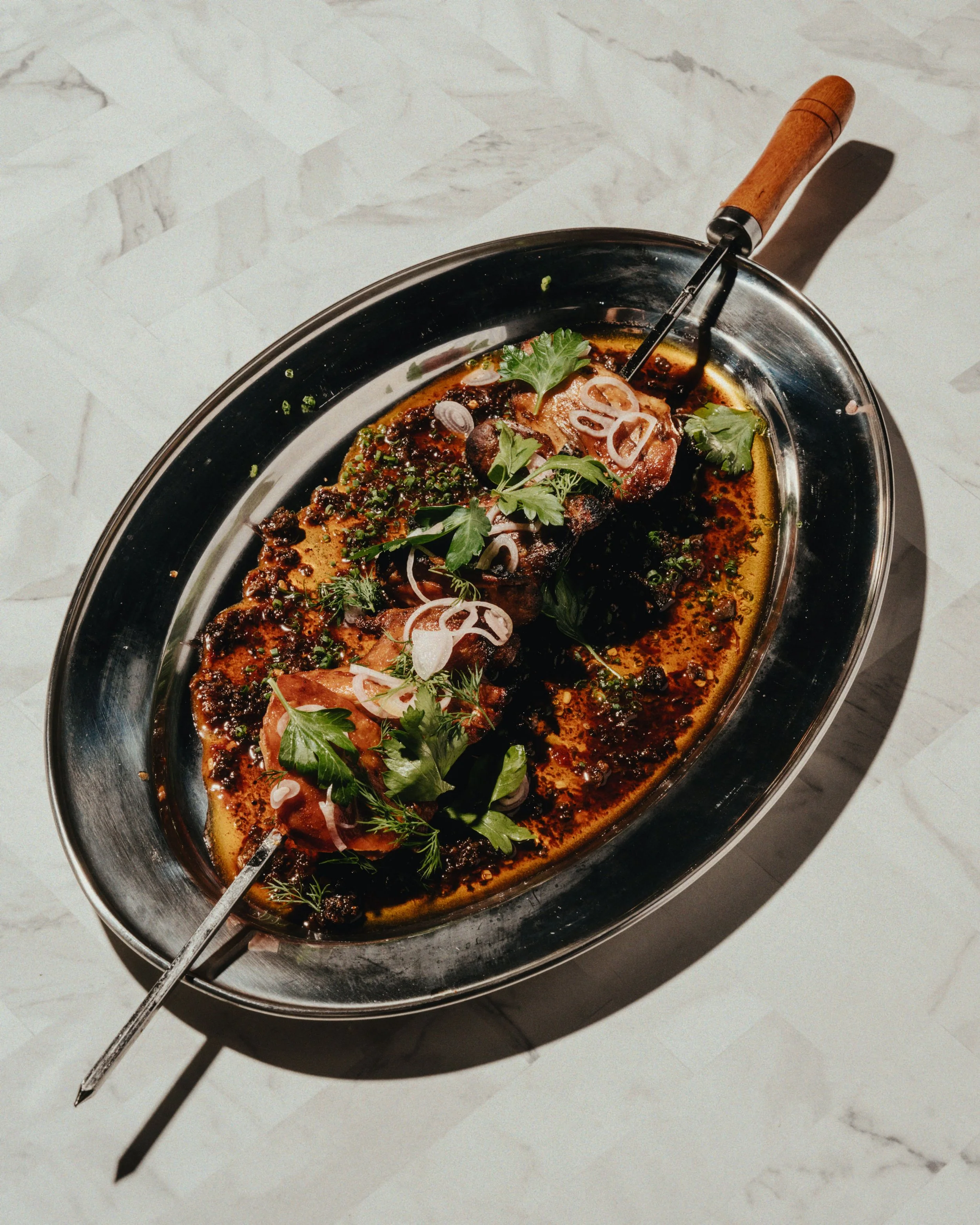 Meat dish garnished with herbs, onions, and sauce served on a silver platter with a wooden-handled utensil.