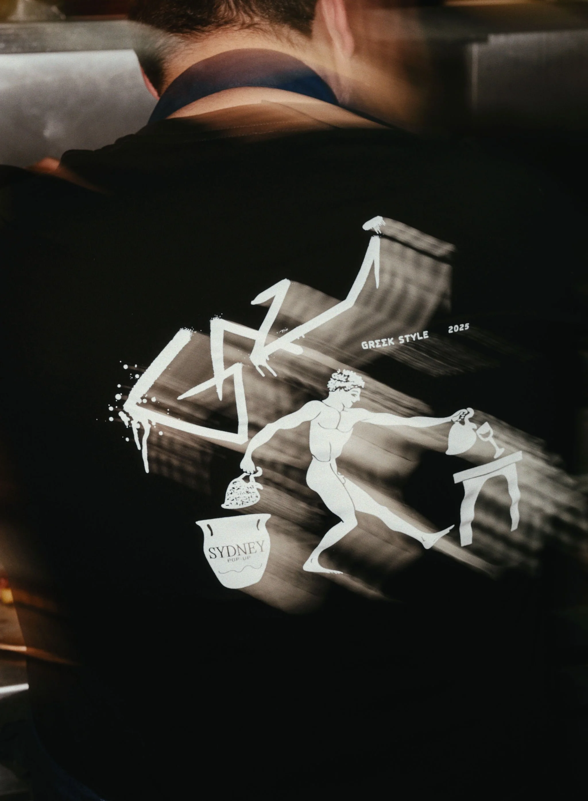 A reflective black surface with a white graphic of a Greek goddess holding two urns, and stylized upward trending arrow graphs, including text that says "GREEK STYLE 2025" and "SYDNEY POP UP."