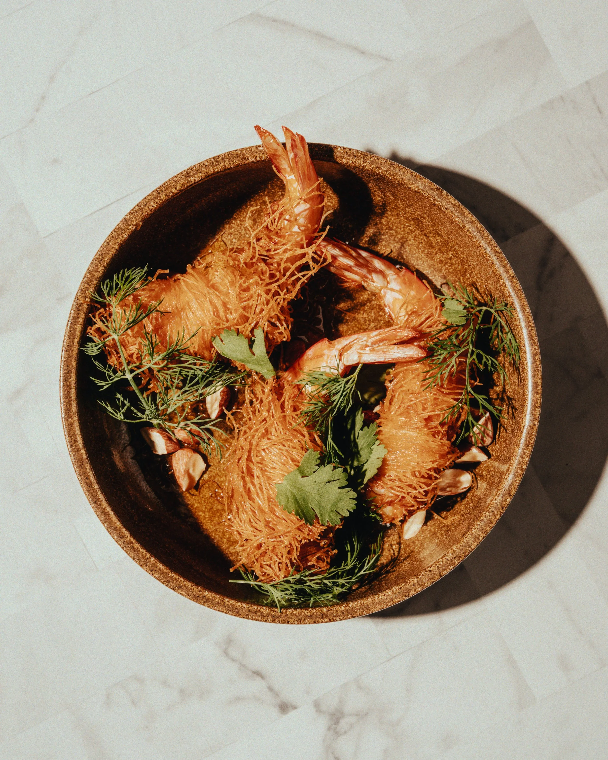 A bowl of cooked shrimp garnished with herbs on a white marble surface.