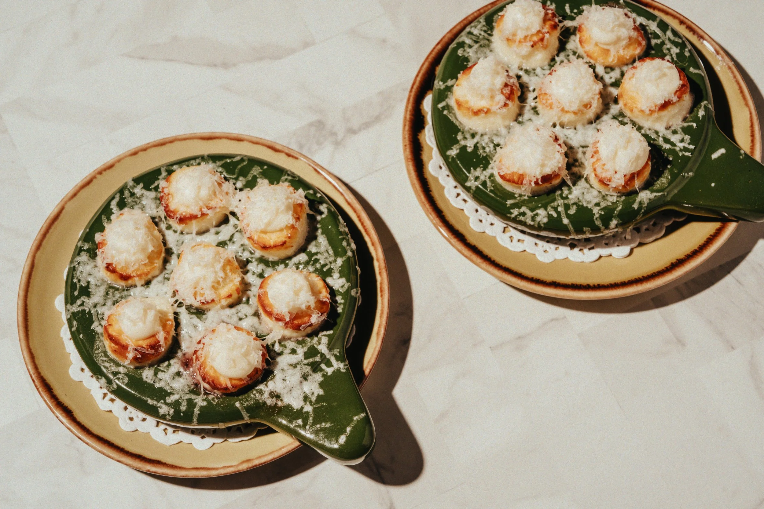 Two ceramic bowls with green and yellow rims, each containing baked Benedict-style eggs topped with cheese, served on white doilies on a white marble surface.
