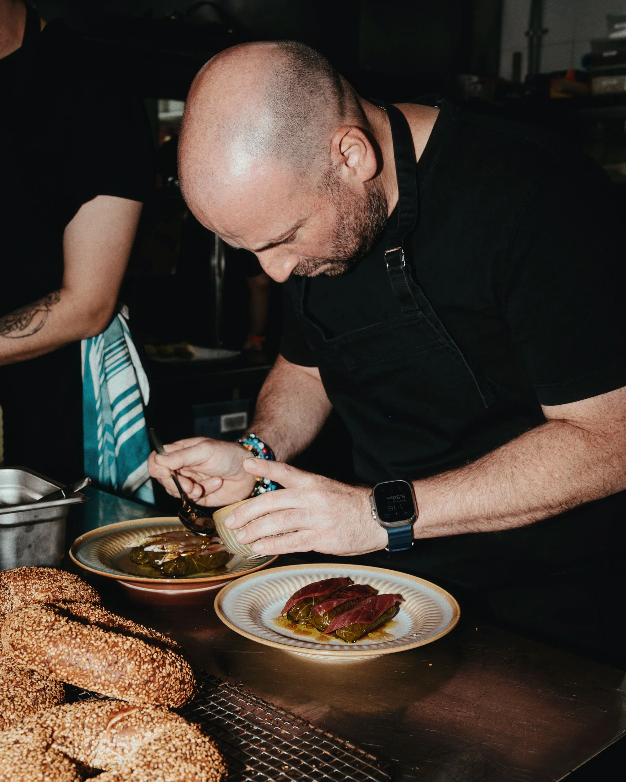 A man with a shaved head and beard, wearing a black shirt, a beaded bracelet, and a smartwatch, prepares and plates stuffed grape leaves in a kitchen. Sesame seed-covered breads are in the foreground.