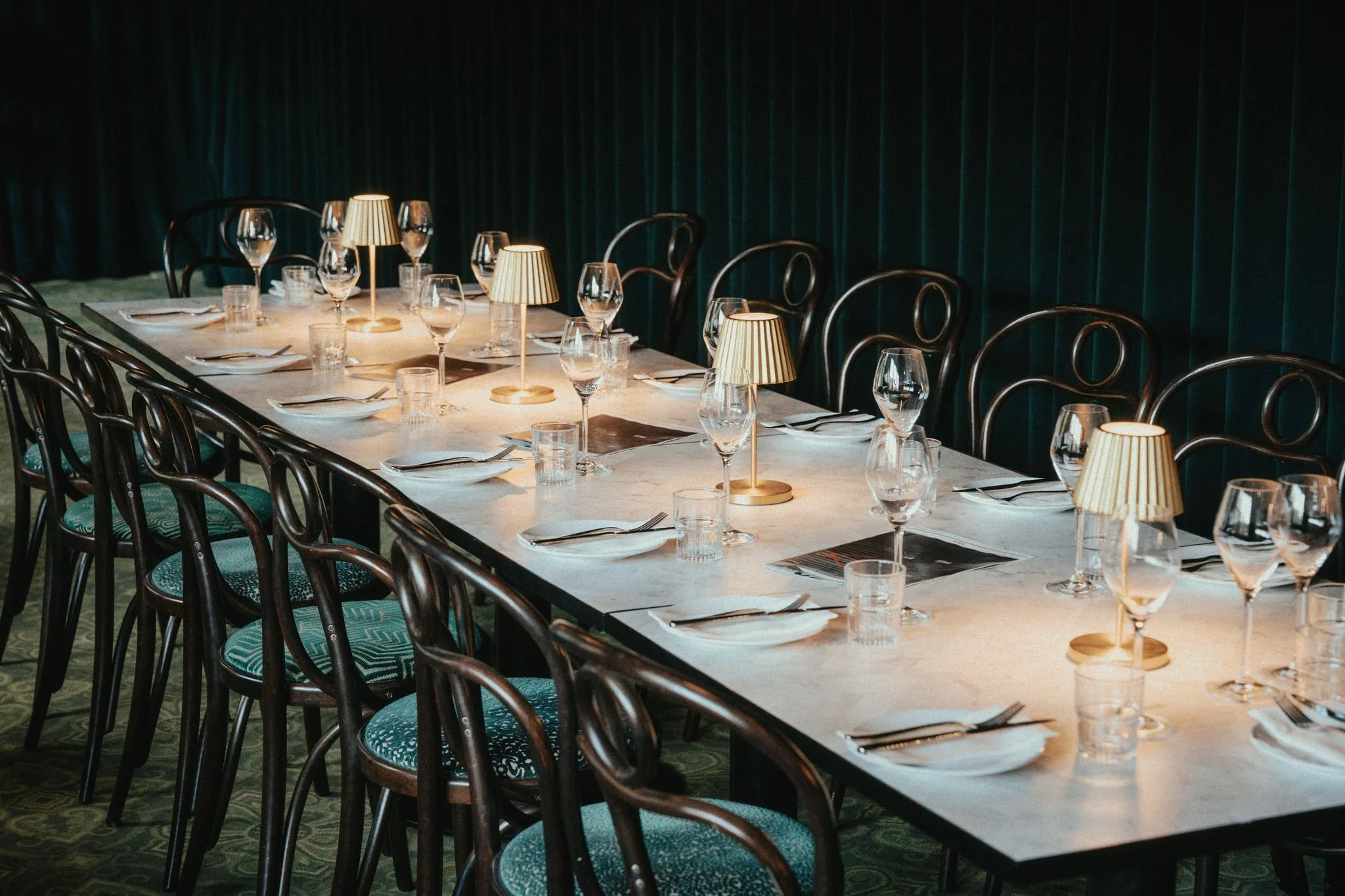 A long dining table set for a formal event with wine glasses, water glasses, plates, silverware, small lamps, and a dark curtain backdrop.