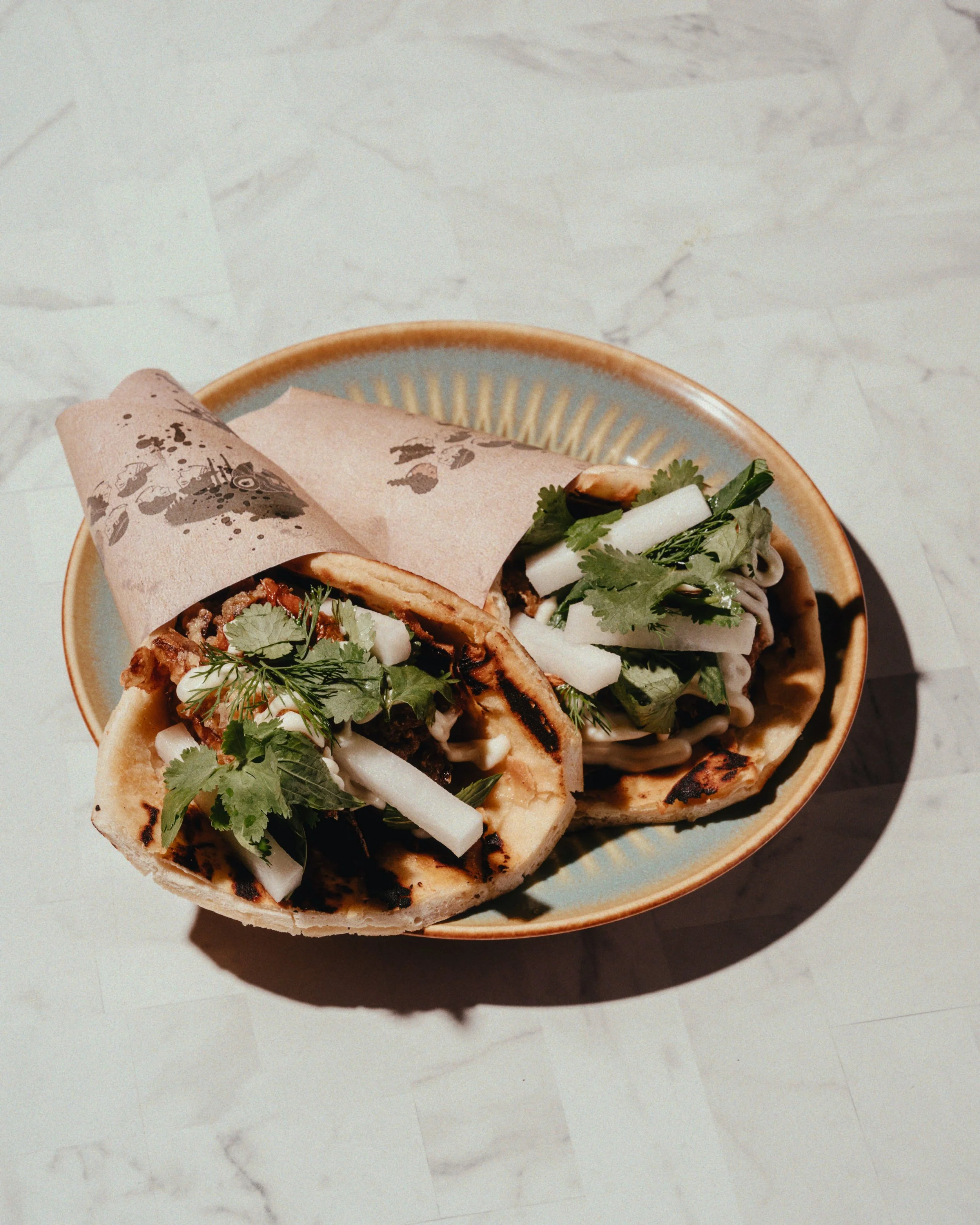 Two grilled tacos filled with meat, topped with cilantro and white onion, served on a ceramic plate on a marble surface.