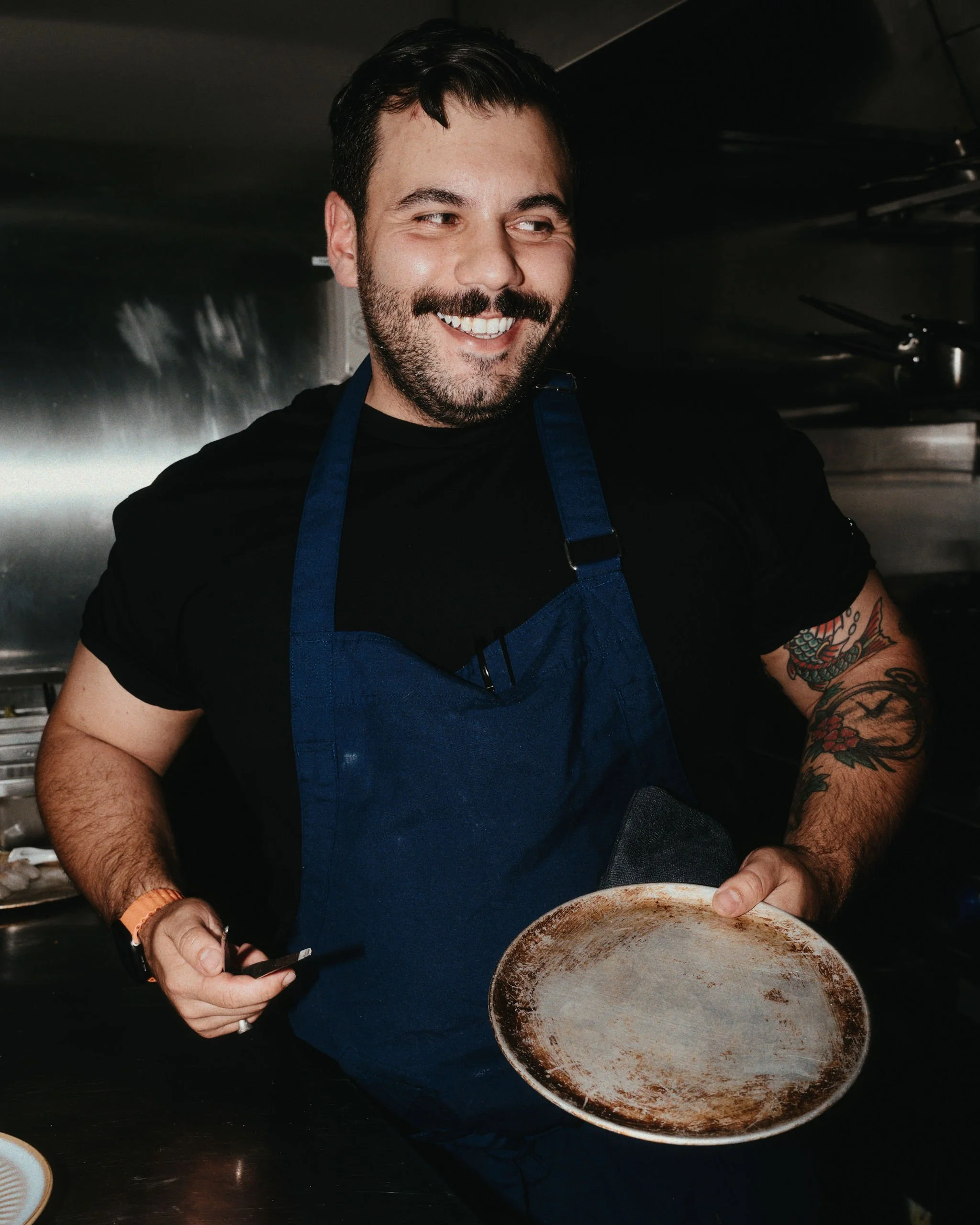 Smiling chef holding a dirty, empty frying pan in a commercial kitchen.