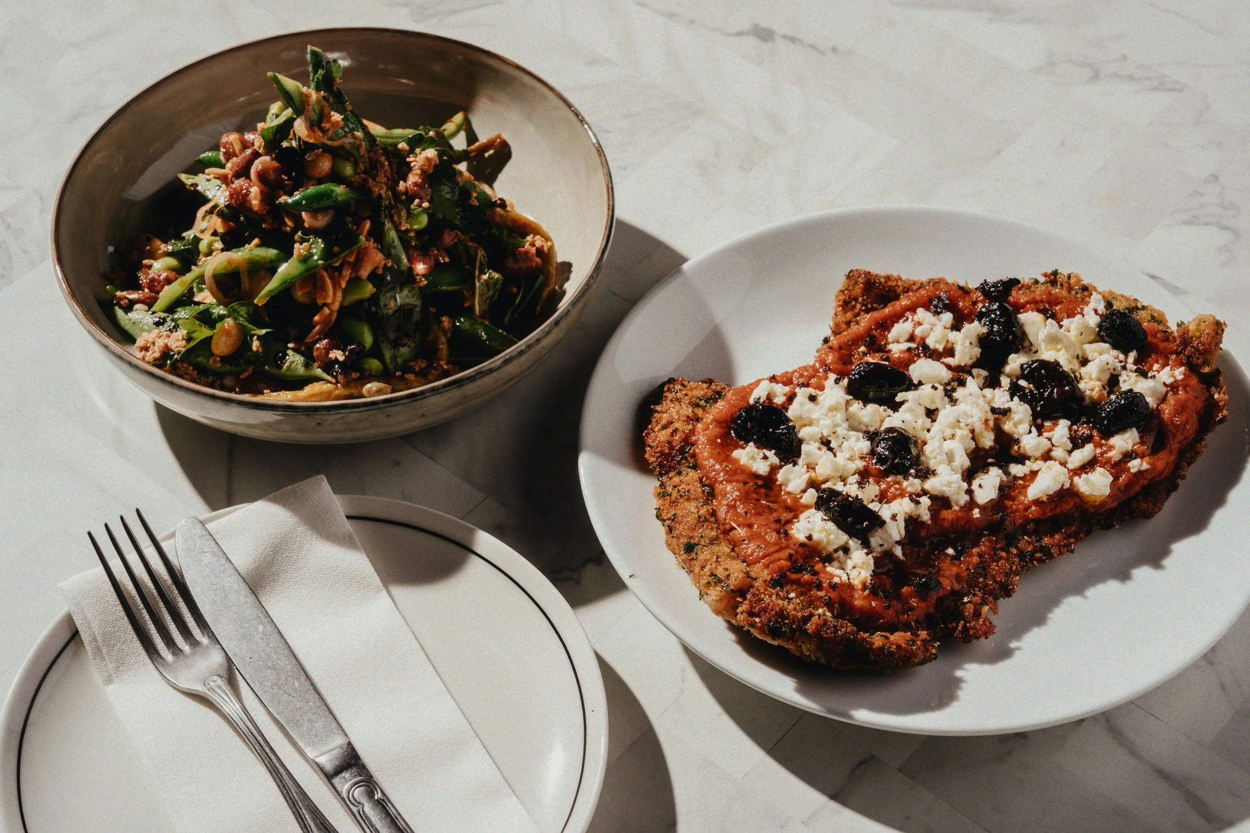 A plate with a piece of breaded baked salmon topped with tomato sauce, crumbled feta cheese, and black olives, a bowl of mixed greens salad with chopped vegetables and seeds, a small empty plate with a knife and fork wrapped in a napkin, all on a lig