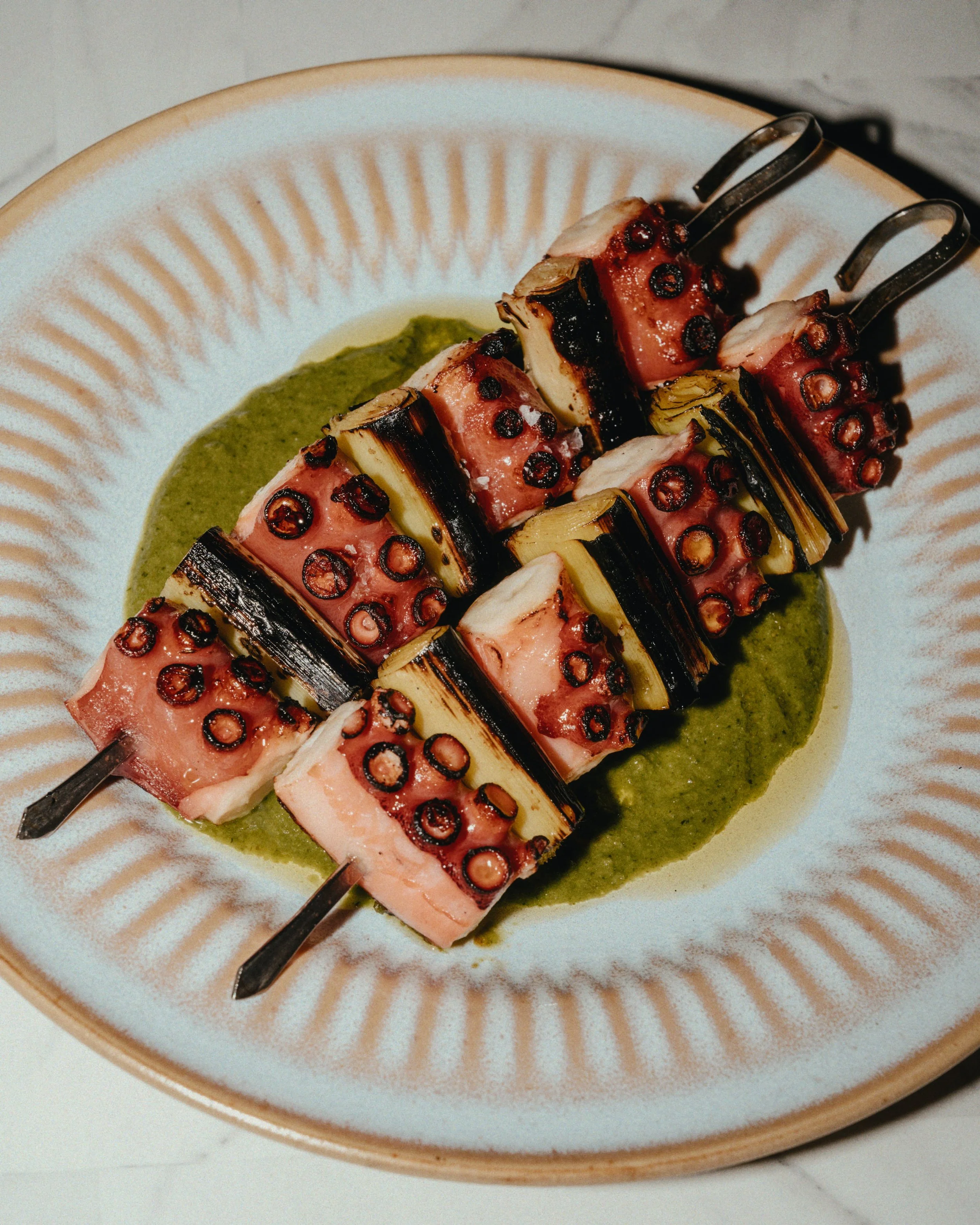 Skewers of grilled octopus, eggplant, and pineapple served on a white plate with green sauce.
