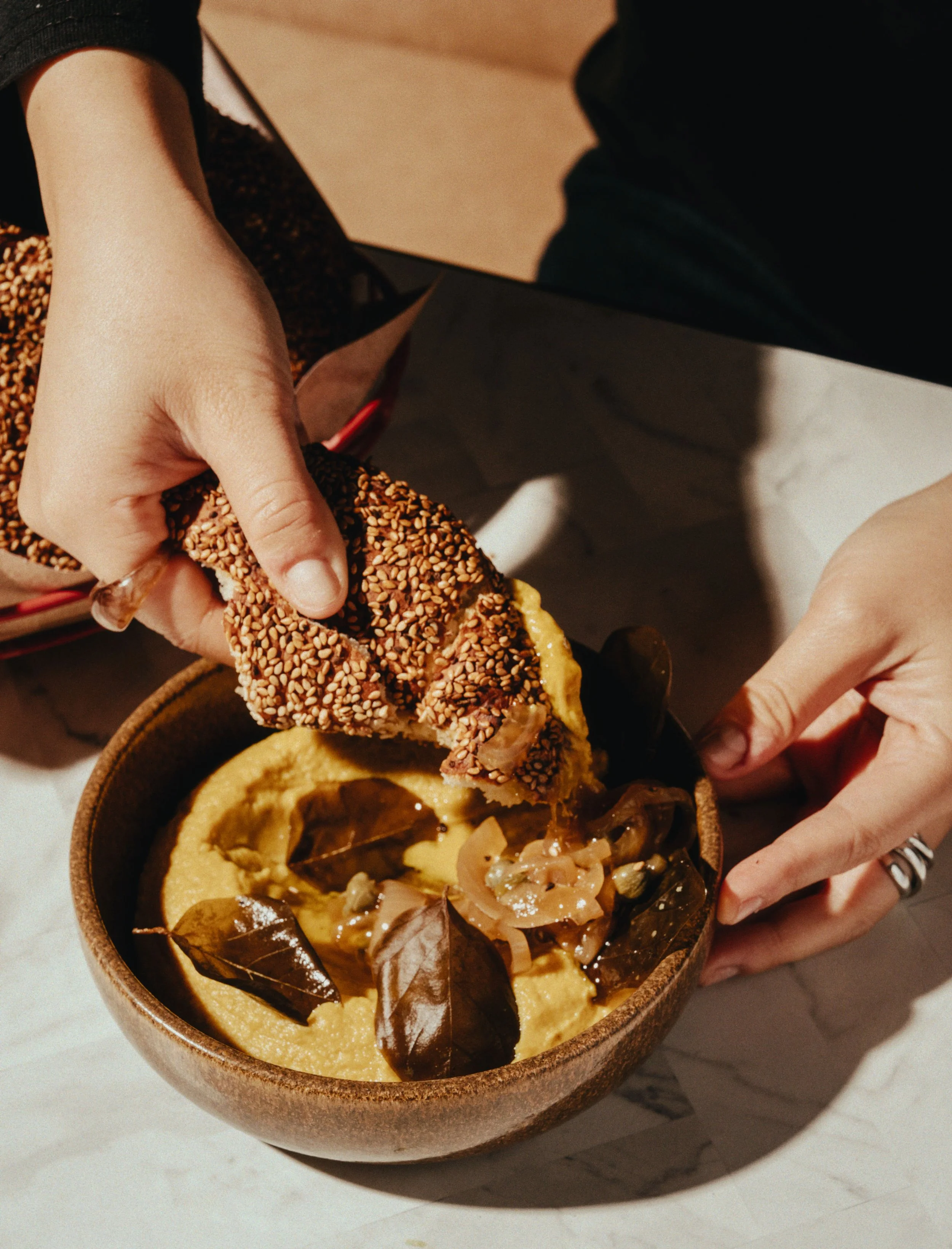 A person dipping a sesame-crusted bread into a bowl of hummus topped with roasted onion, bay leaves, and olive oil on a marble surface.