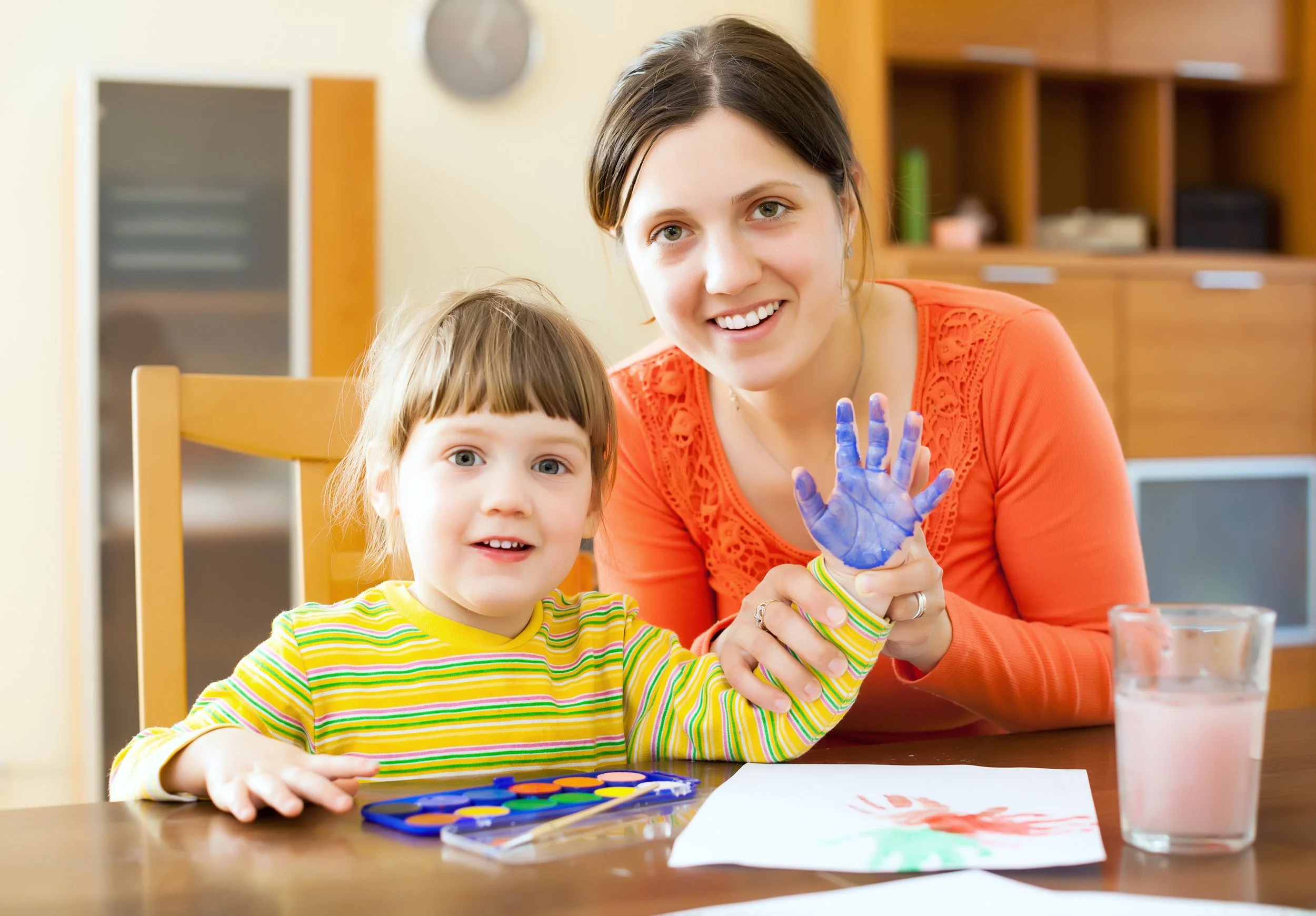 A woman with brown hair and green eyes smiles as she shows her painted hand to a young girl with brown hair and blue eyes, sitting at a table with paint supplies, in a room with wooden shelves and a beige wall.