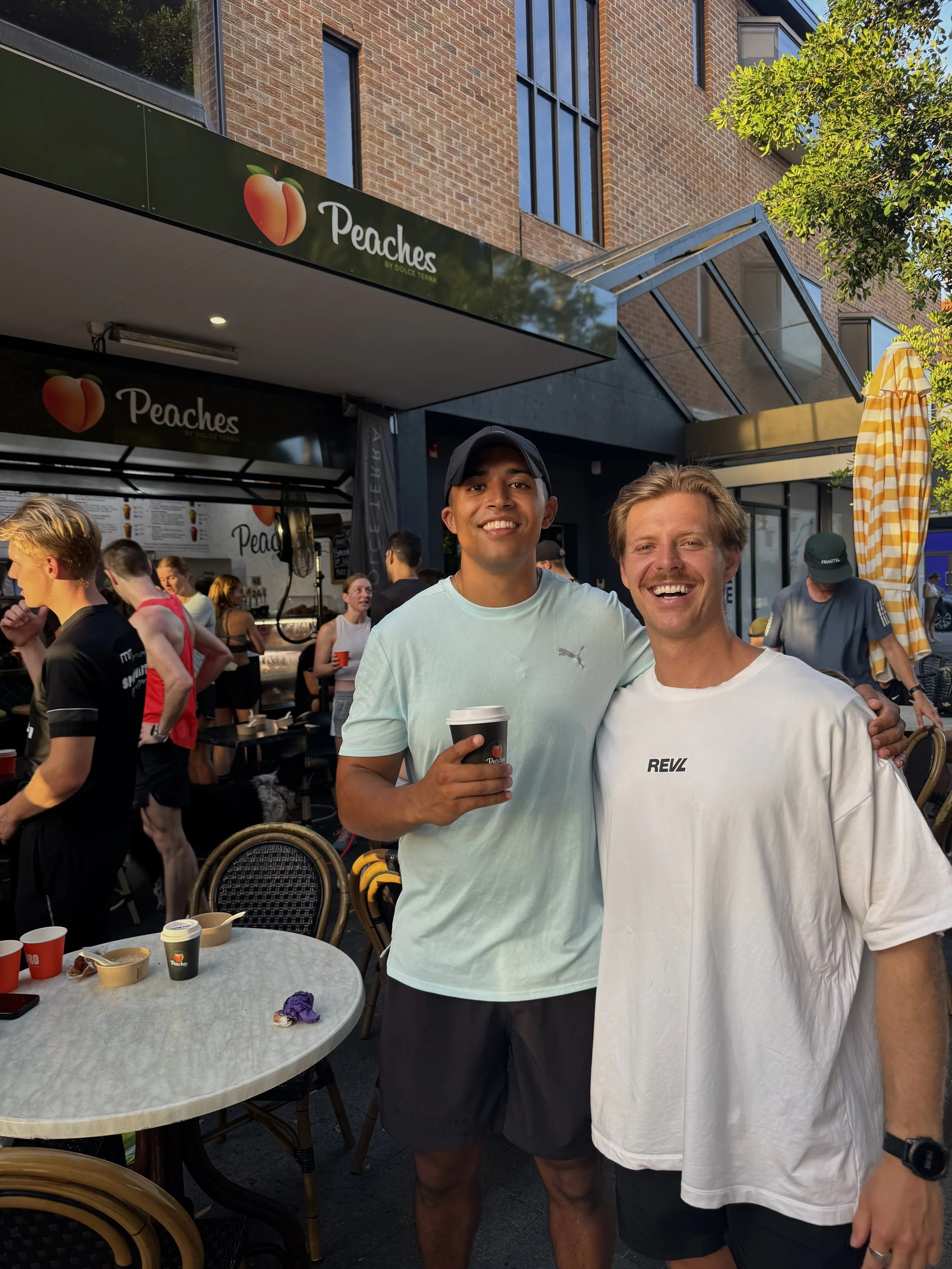 Two smiling men standing outdoors in front of a restaurant named 'Peaches,' with people in the background, one holding a coffee cup.