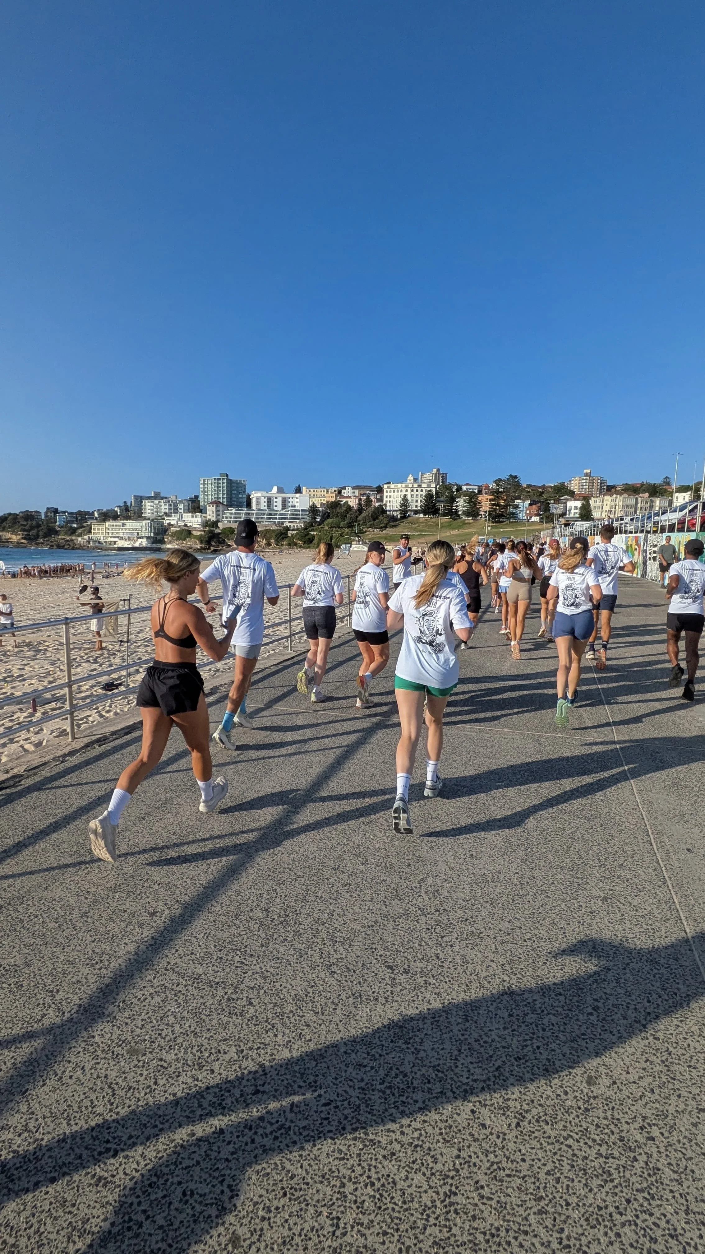 Group of people running on a promenade near a beach with urban buildings in the background on a sunny day.