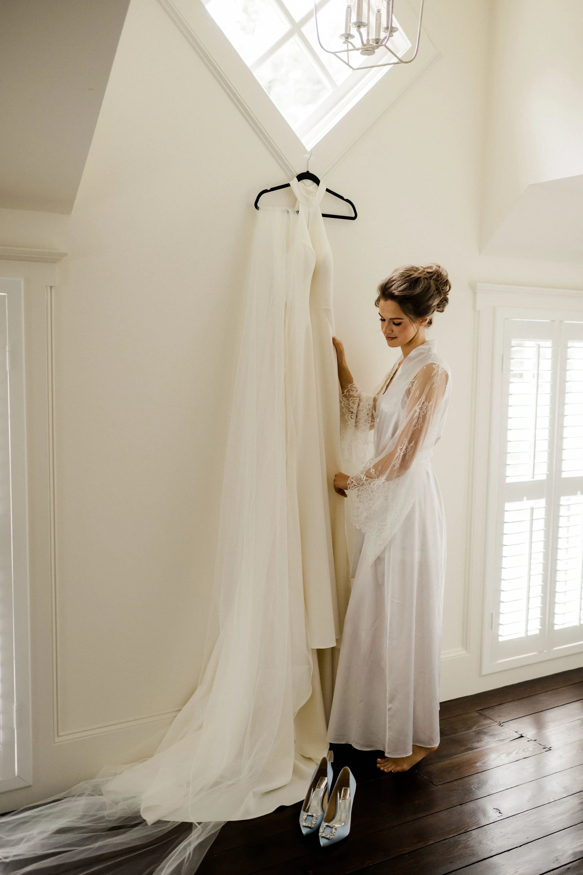 A woman in a satin robe standing barefoot beside a hanging wedding dress with a long train and veil, in a bright room with wooden floors and white walls.