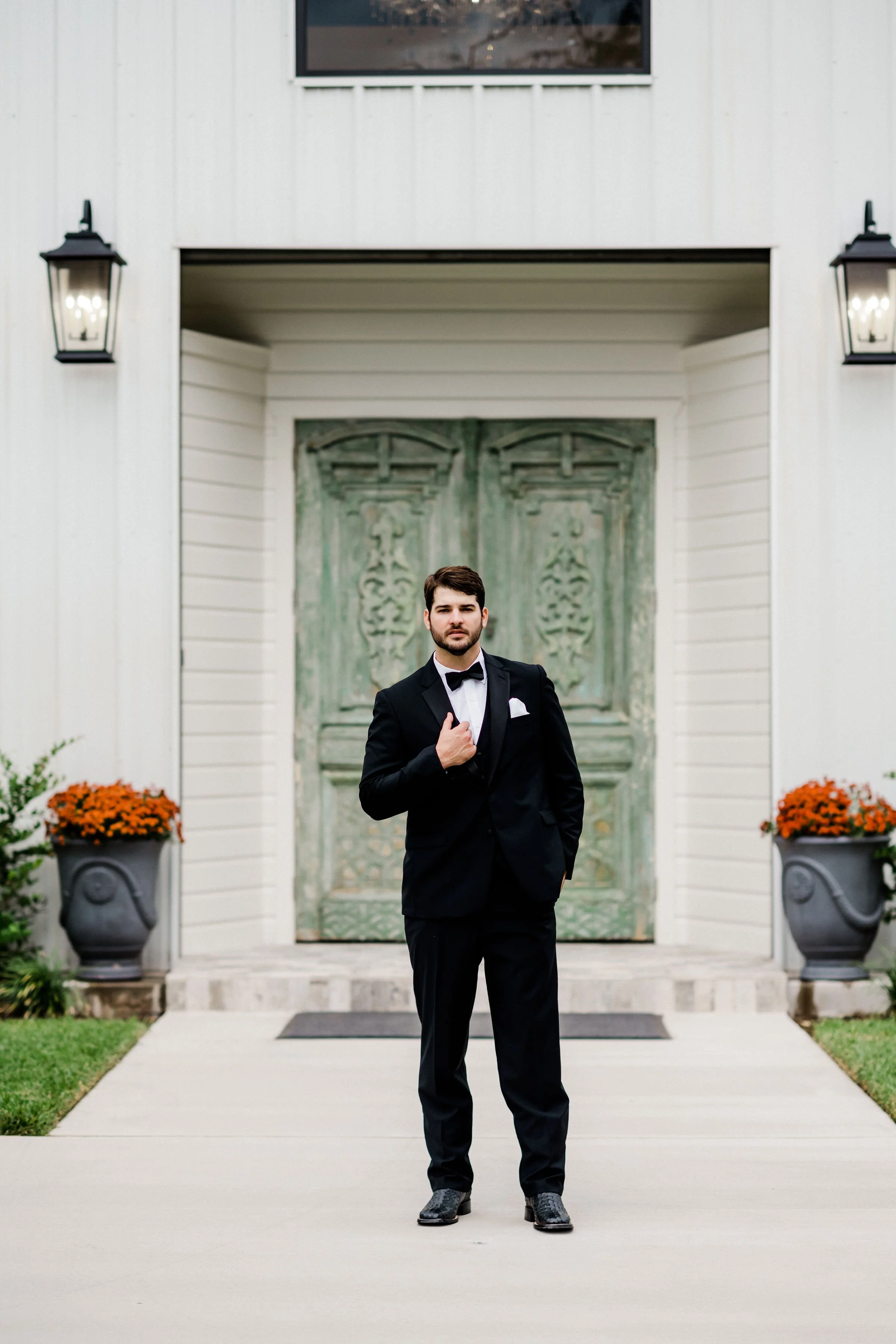 A man in a black tuxedo with a bow tie standing outside in front of a house with green double doors and two outdoor wall lamps, flanked by large potted orange flowers.