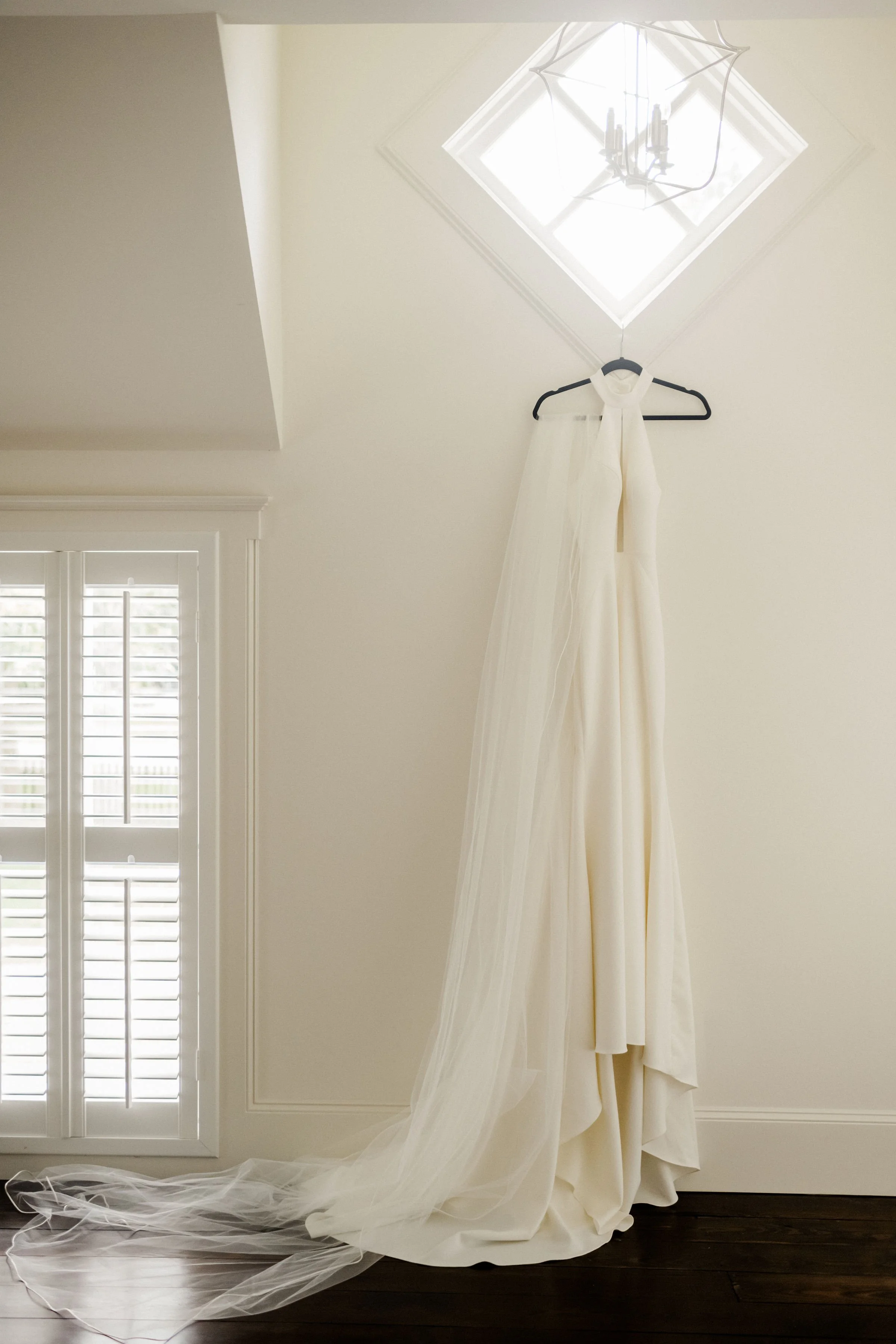Wedding dress hanging on a black hanger against a white wall, next to a window with white shutters, with a ceiling light fixture above.