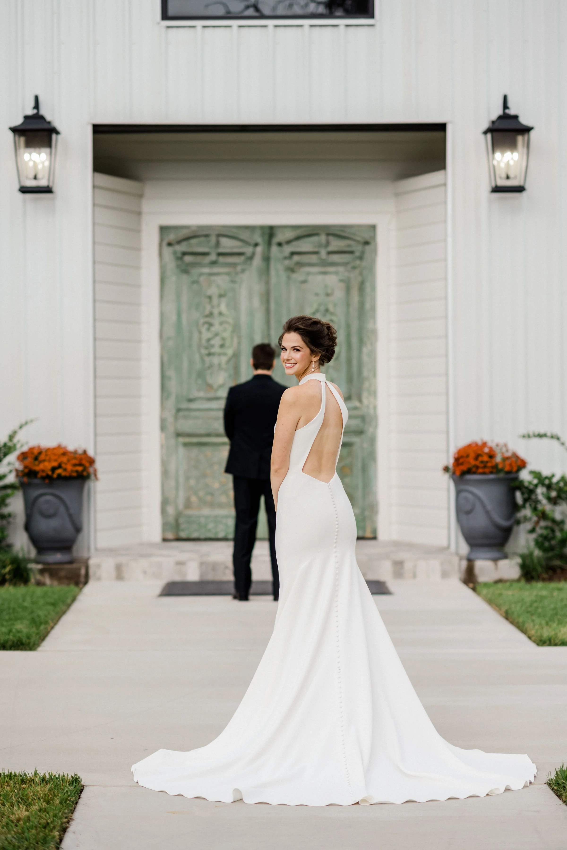 Bride in a white wedding dress smiling and turning towards camera, groom in a black suit facing away in front of a green door outside a white building with lanterns and orange flowers.