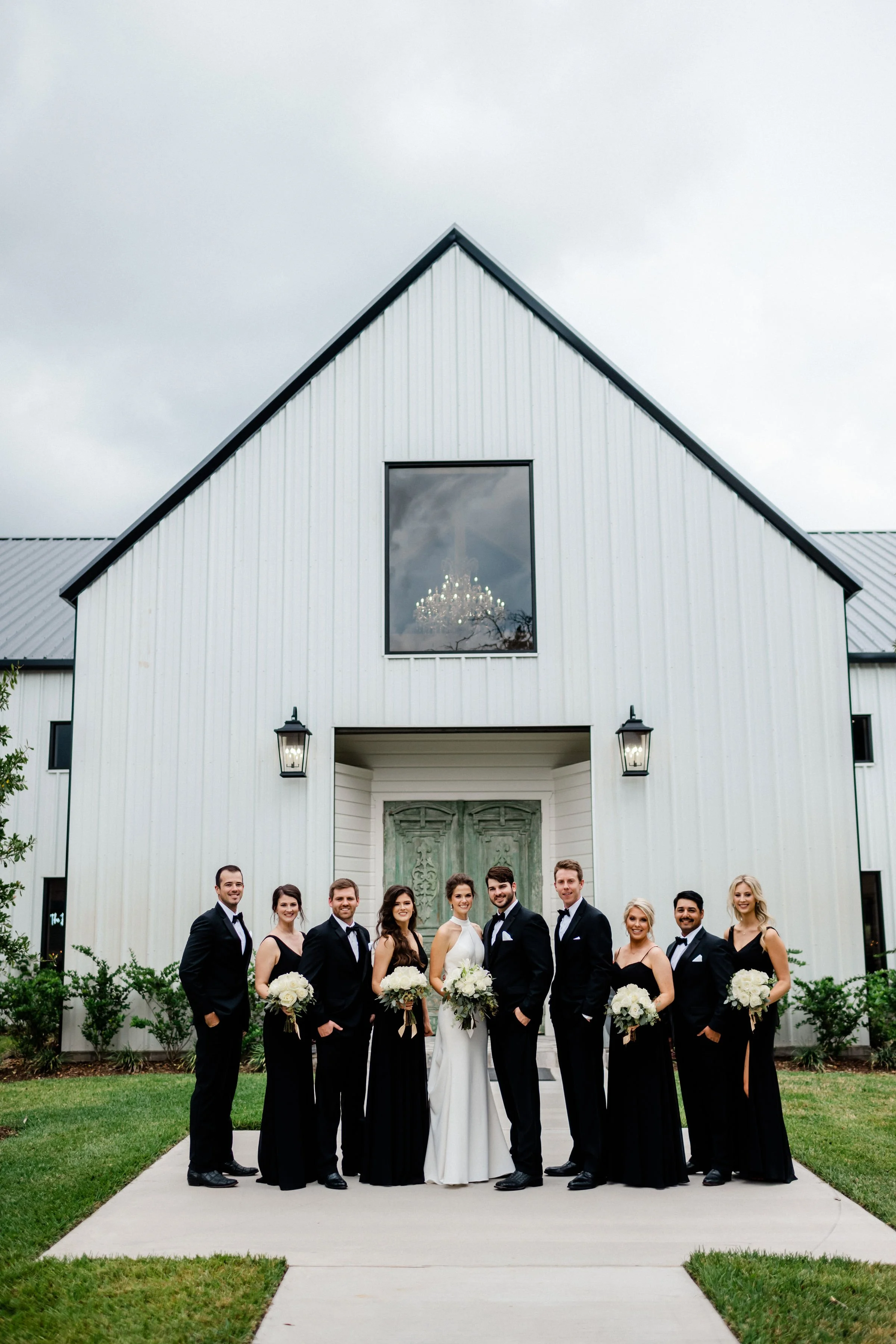 A wedding party standing outside a modern white chapel with green doors, including a bride and groom surrounded by bridesmaids and groomsmen, all dressed in formal attire and holding bouquets of white flowers.