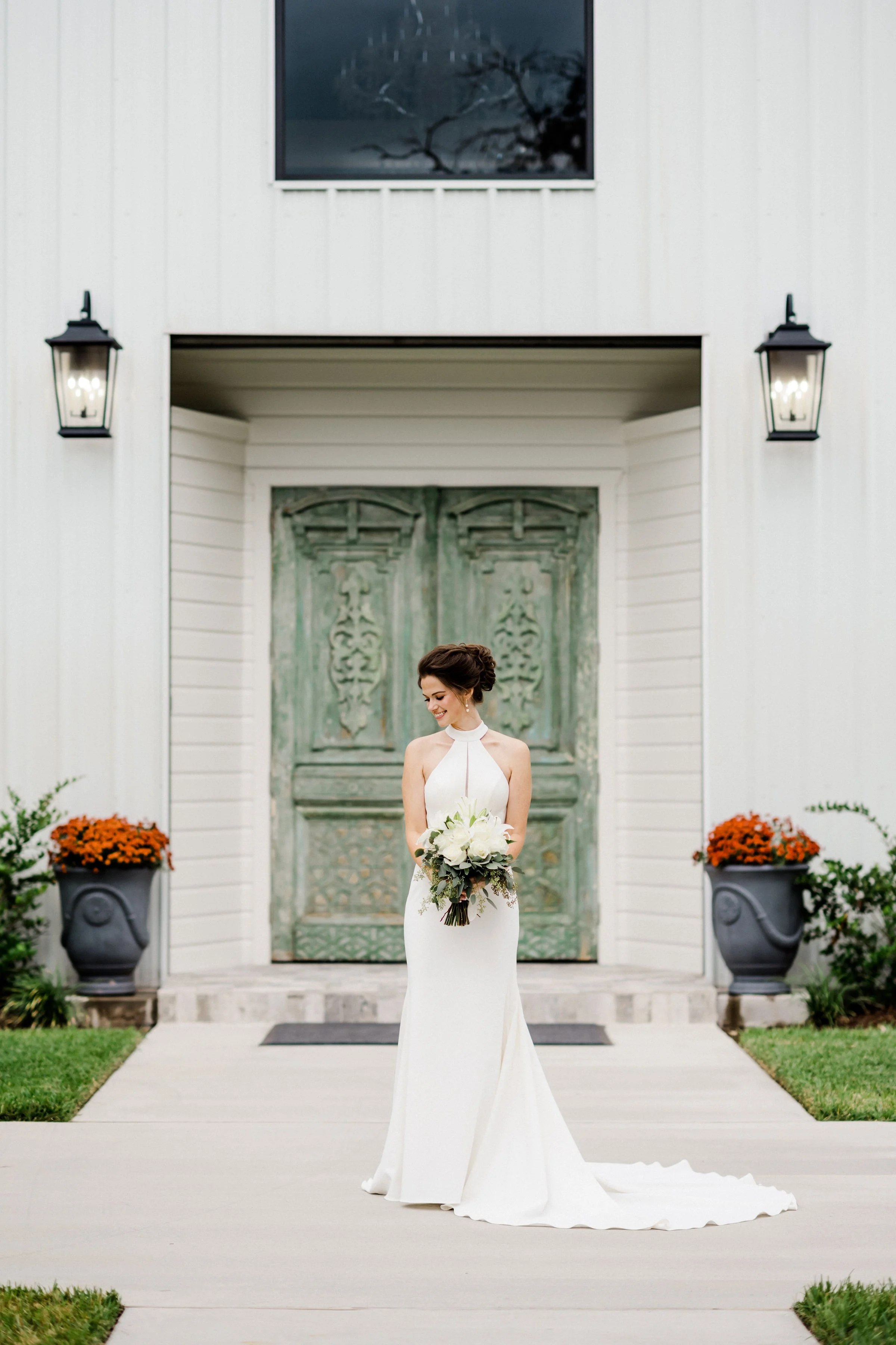 Bride in a white gown holding a bouquet in front of a rustic wooden door with potted orange flowers on either side.