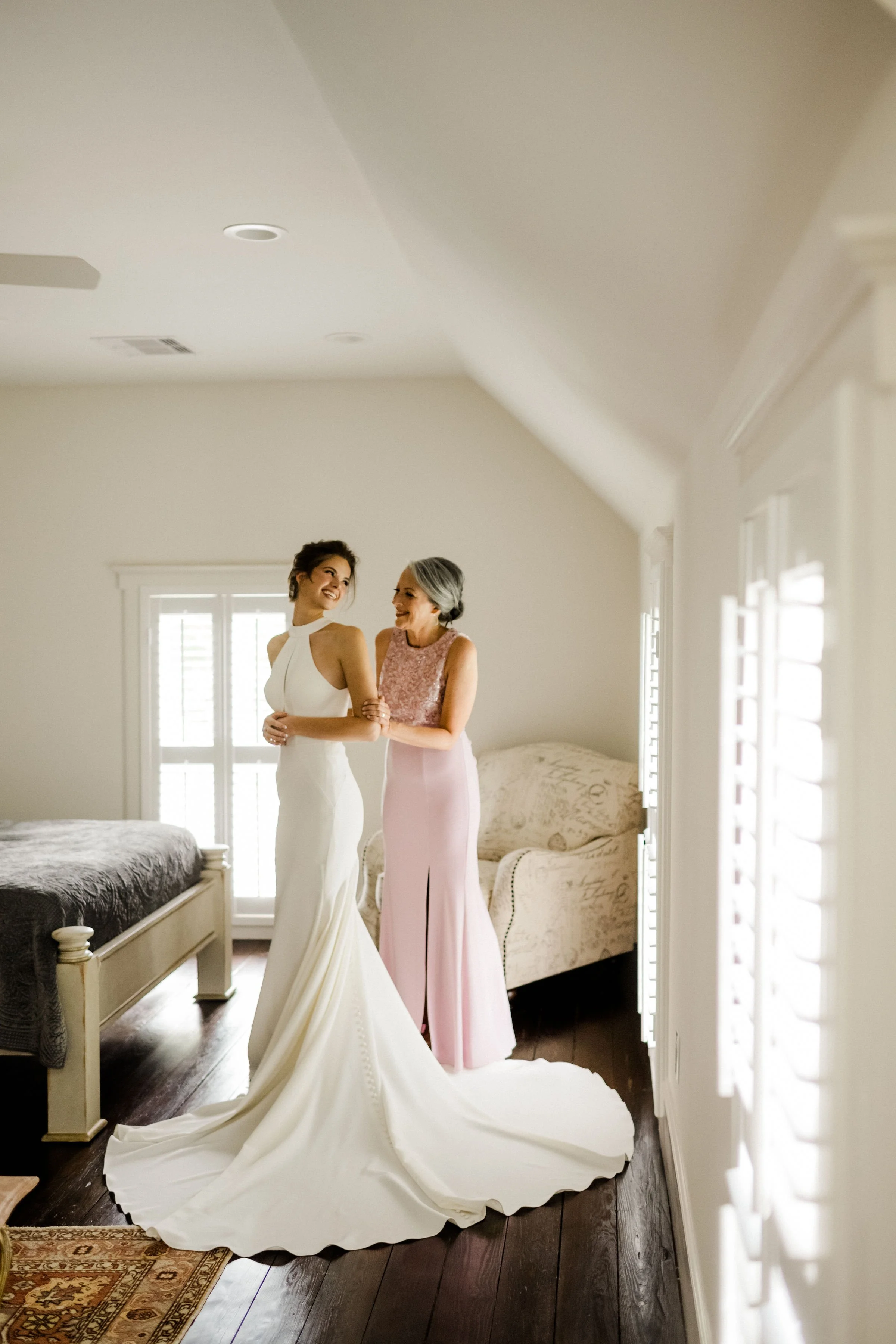 A bride in a white wedding gown standing with an older woman in a pink dress in a bright bedroom.