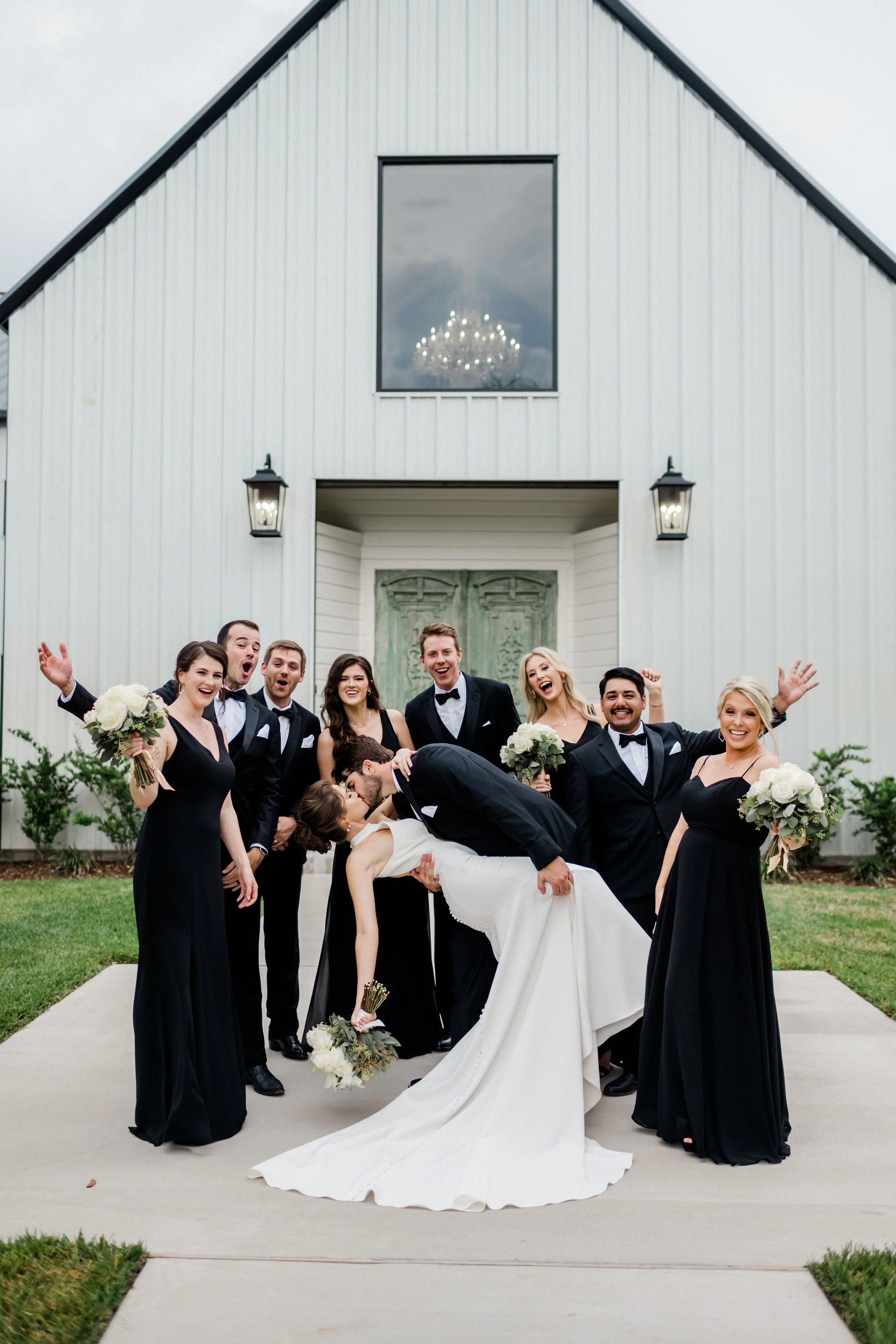 A group of wedding party members, including the bride and groom, posing in front of a white barn-style building. The bride is wearing a white wedding dress and is being dipped by the groom, who is in a black tuxedo with a bow tie. The bridal party is