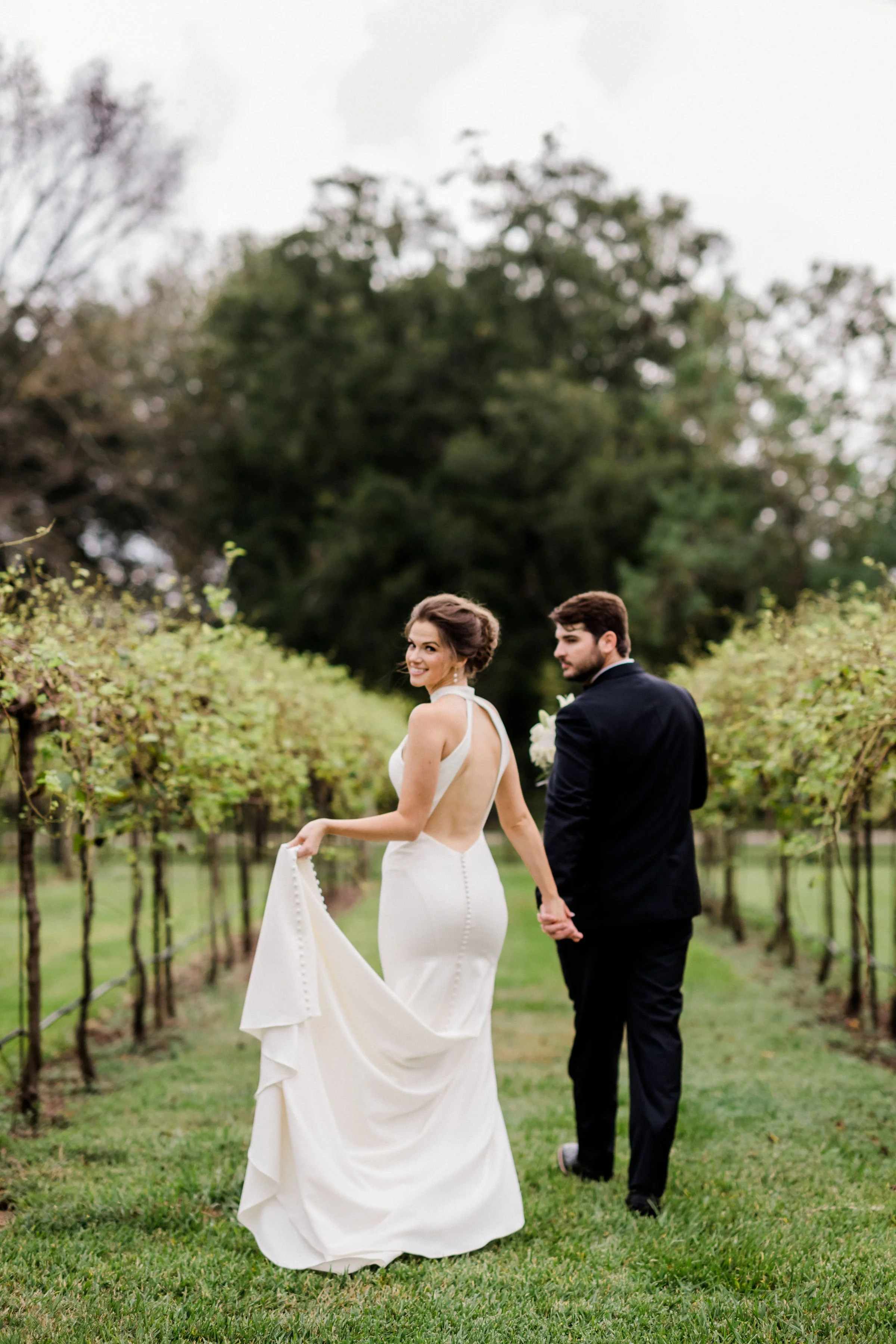 A bride and groom holding hands in a vineyard on their wedding day, with the bride looking back and smiling at the camera.