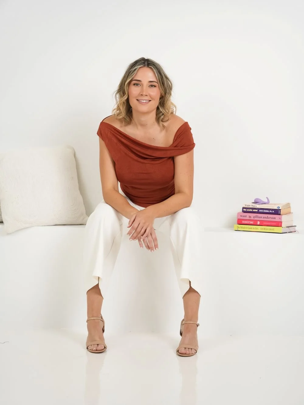 A woman sitting on a white bench, smiling, wearing a rust-colored off-shoulder top and white pants, with a stack of books beside her.