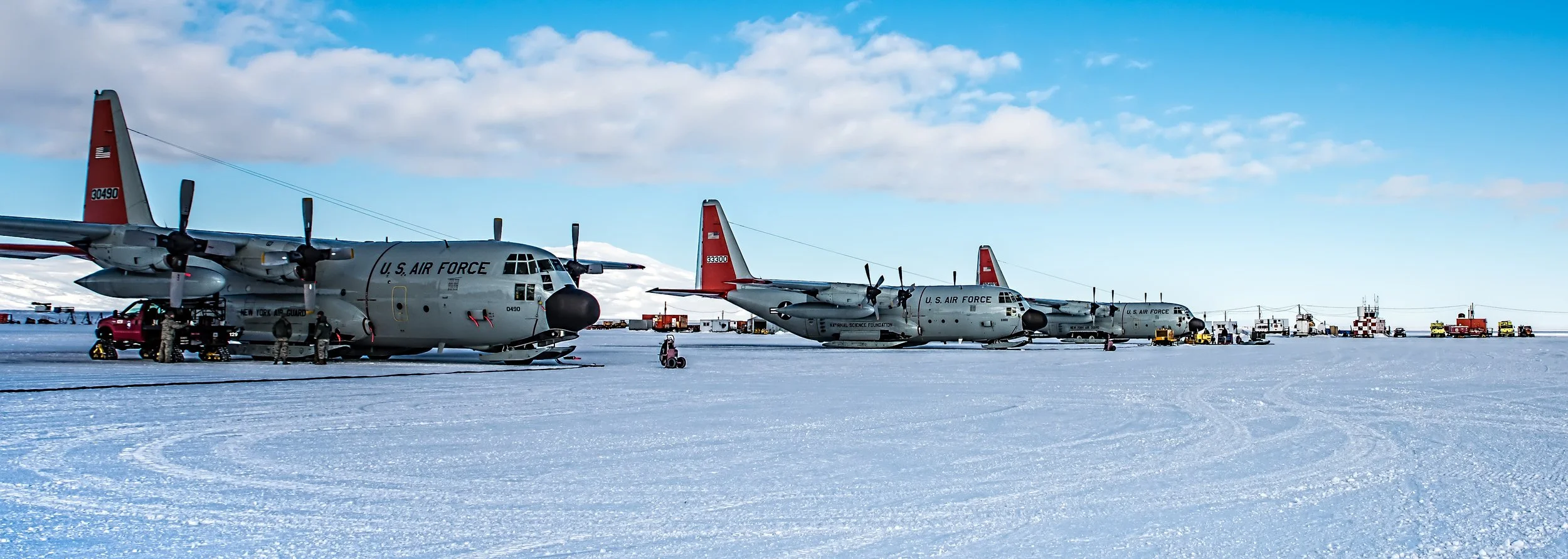 U.S. Air Force C-130 aircraft parked on icy ground with a clear blue sky, snow-covered landscape, and support vehicles nearby.