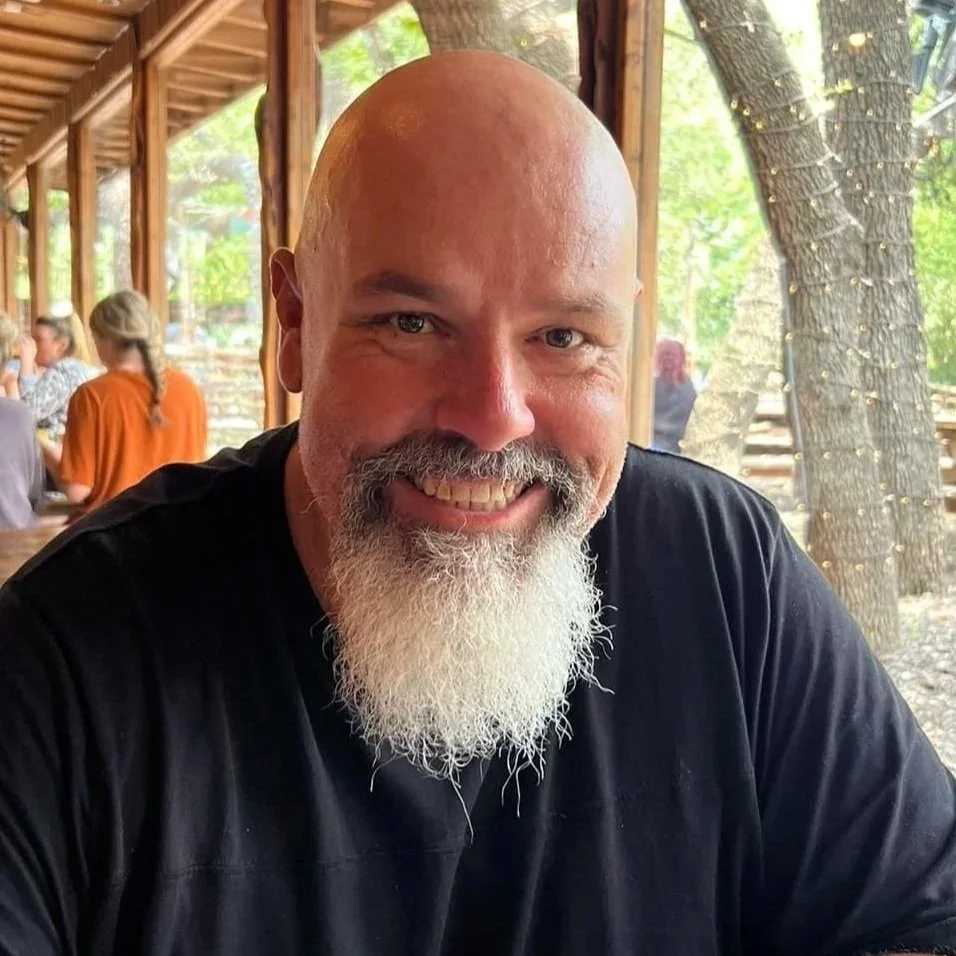 Nathan Baxter, who is the smiling bald man with a white beard sitting at an outdoor wooden restaurant or cafe, with other people in the background and trees outside.