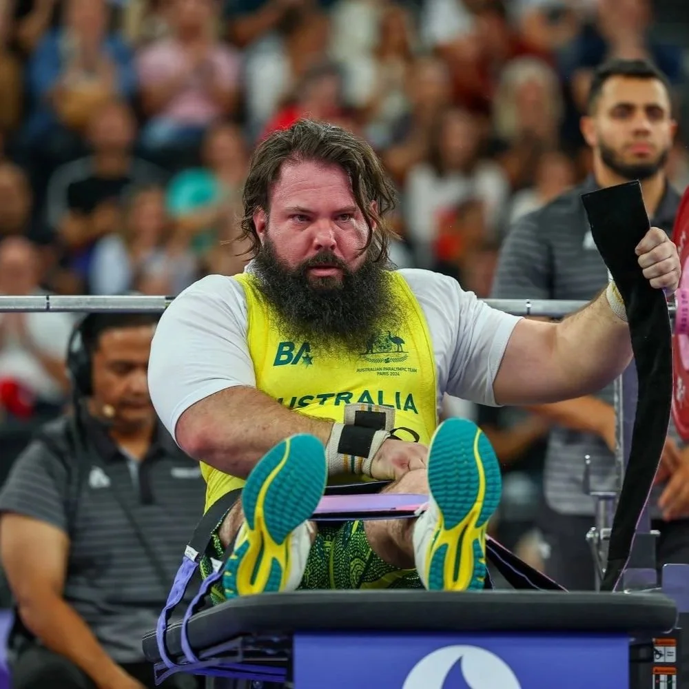 Ben Wright, a male Paralympic athlete with a beard and long hair, wearing a yellow and white shirt and colourful sneakers, participating in parapowerlifting and is about to bench press at the Paralympic Games, surrounded by spectators and officials.