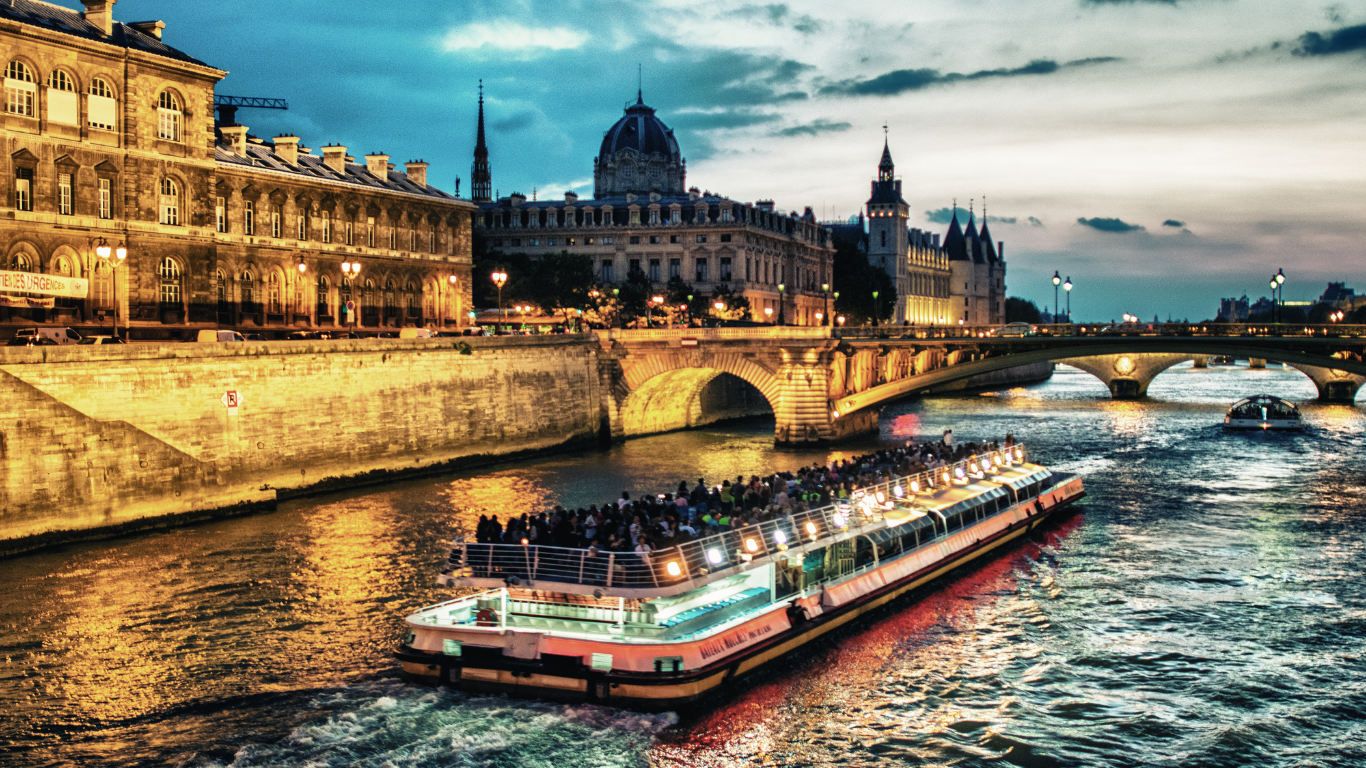 Scenic view of a river in Paris with a tourist boat cruising, historic buildings, and a bridge at dusk.