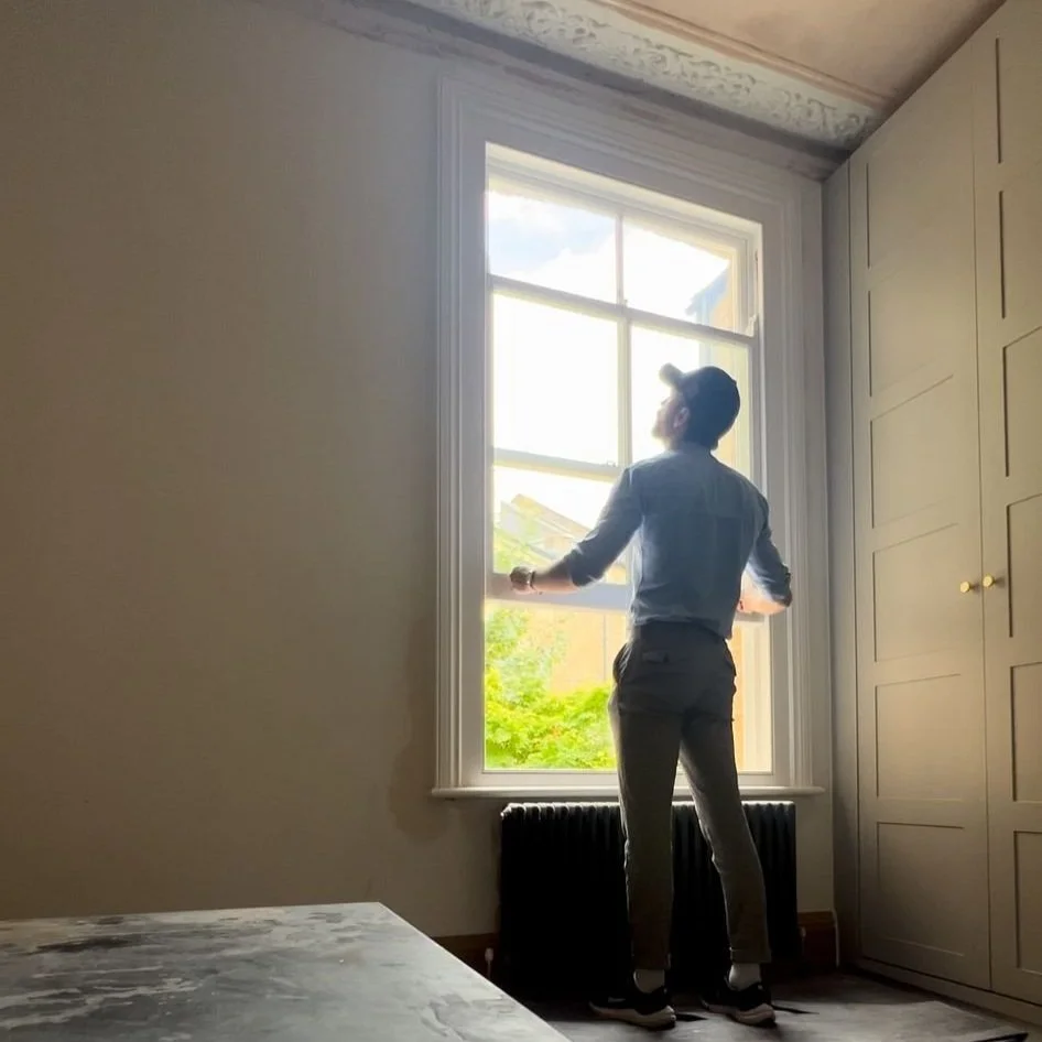 Person standing by a window in a room with minimal decoration, wearing casual clothes and a cap, looking outside.