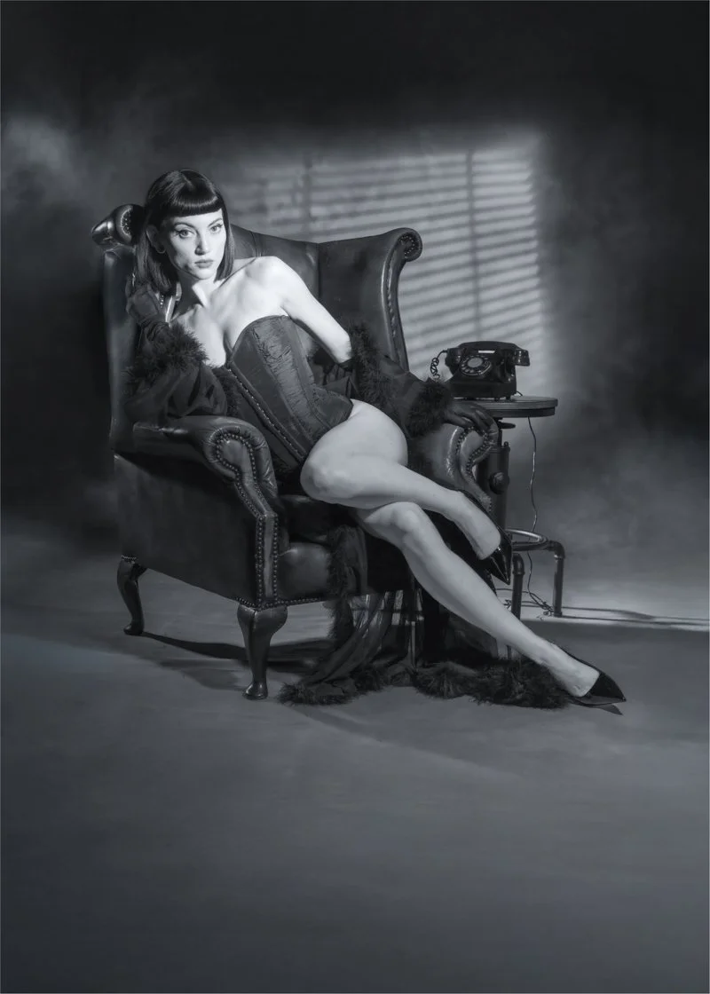 Jordan in vintage corset poses in a wingback chair with dramatic noir lighting and an old rotary phone beside her. Photograph by Andy Shaw Photography