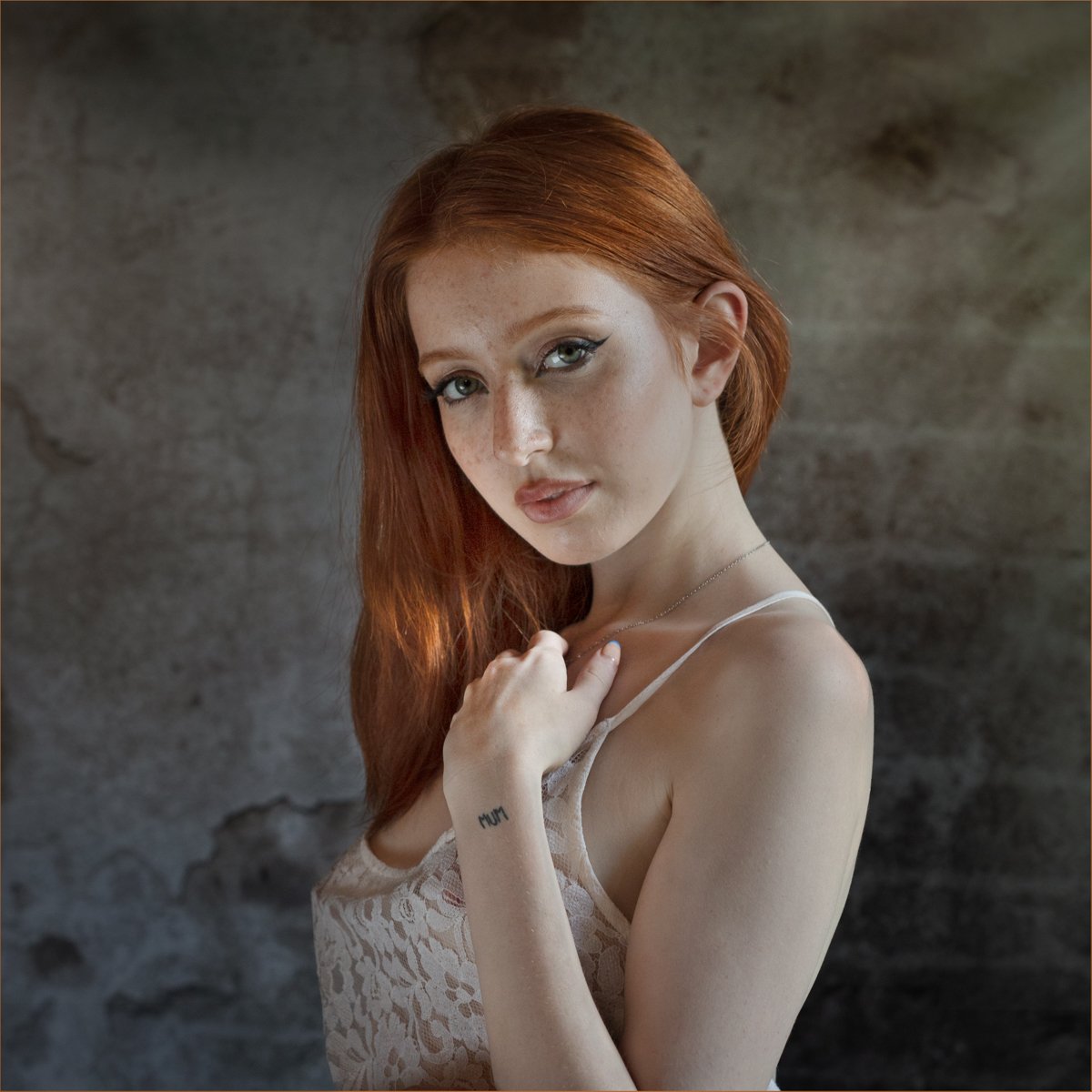 Young woman with long red hair and lace top poses with an over-the-shoulder look against a textured wall. Photograph by Andy Shaw