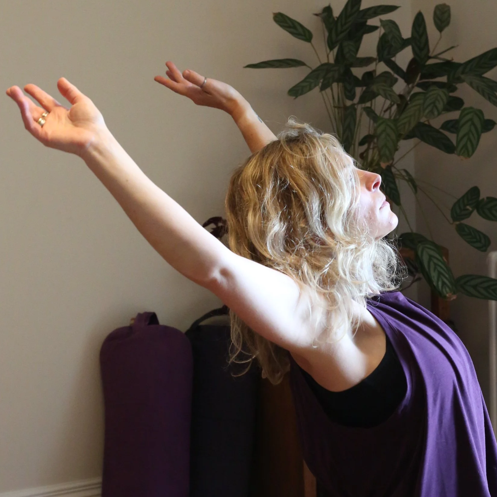 A woman with blonde, curly hair practicing yoga indoors with her arms extended upward and her head tilted back, sitting on a purple cushion, in front of a green leafy plant and beige wall.
