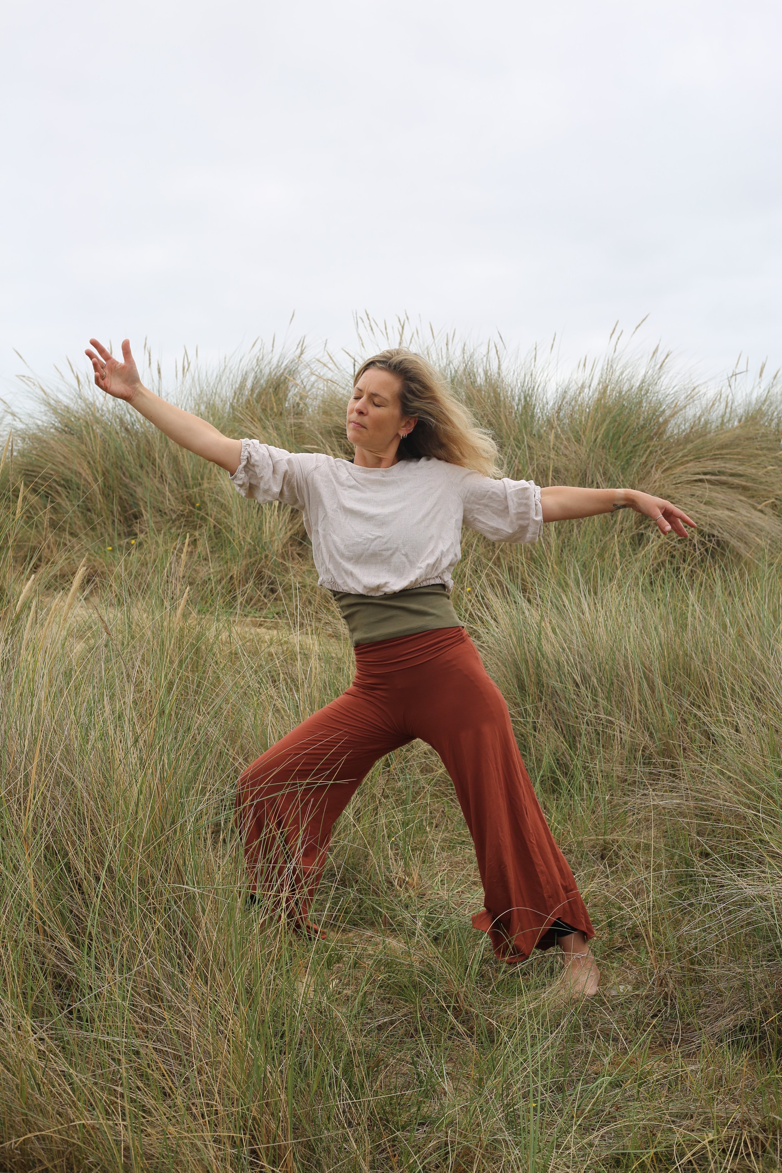 A woman with long hair practicing yoga outdoors in a grassy field with tall grass and a cloudy sky.