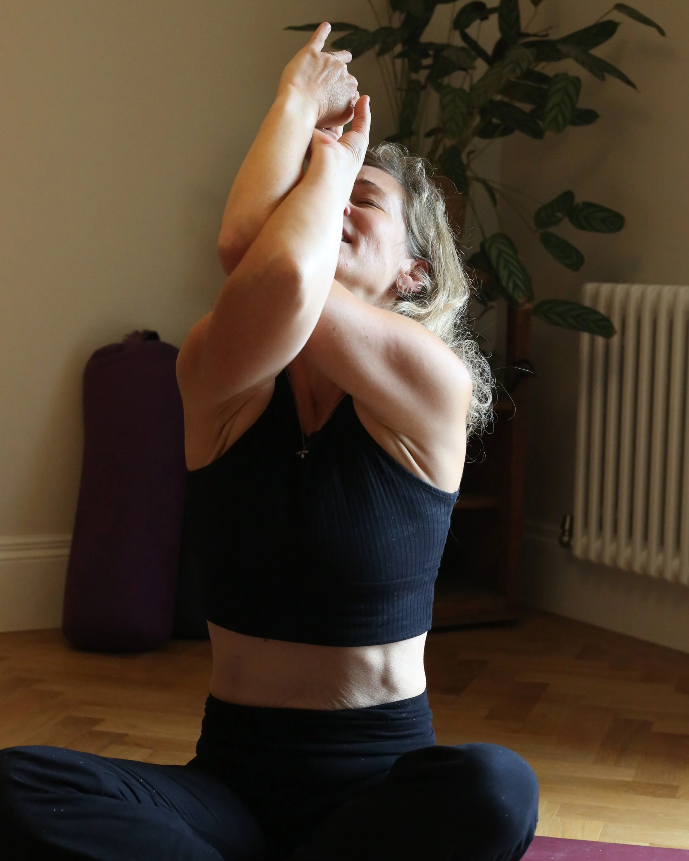 A woman engaging in a yoga pose indoors, sitting cross-legged on a yoga mat, with her arms raised and hands pointing upward, smiling and eyes closed.