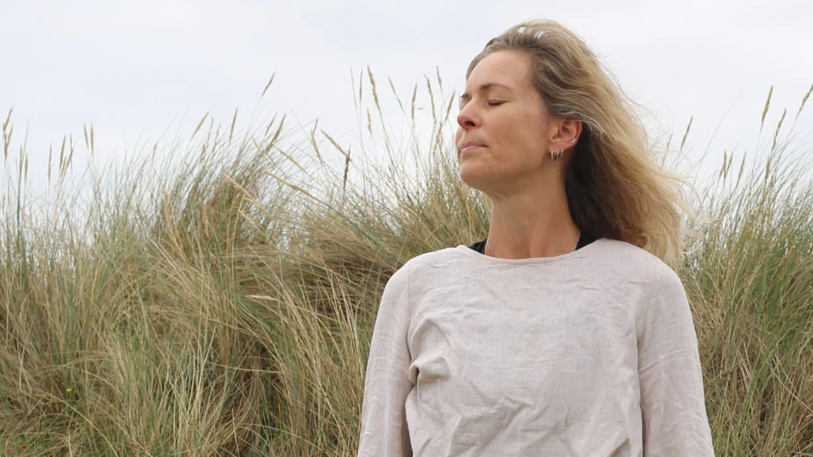 Woman with closed eyes standing outdoors in front of tall grasses, wind blowing through her hair.