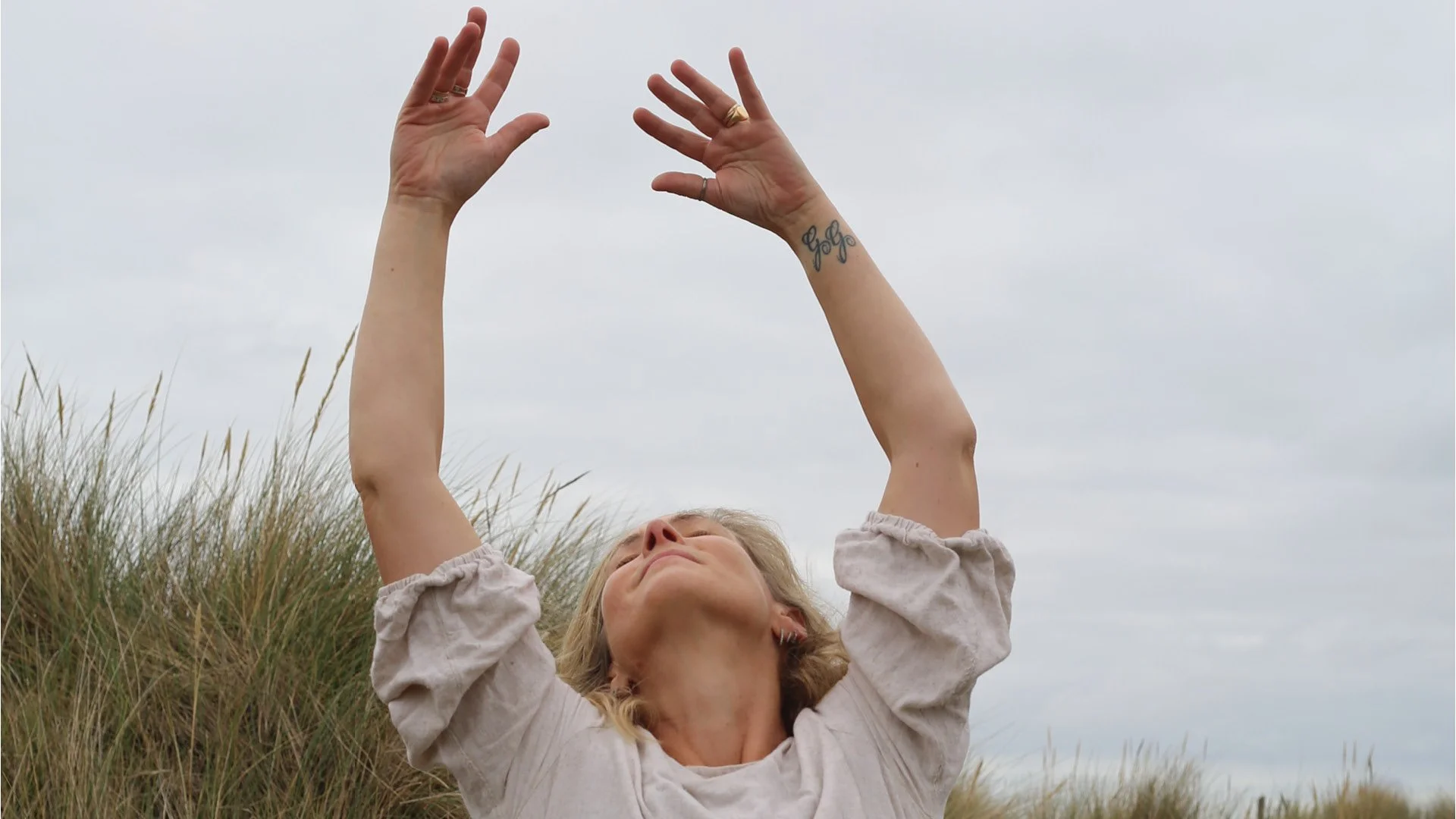 A woman raising her arms above her head while lying in tall grass outdoors, with cloudy sky in the background.