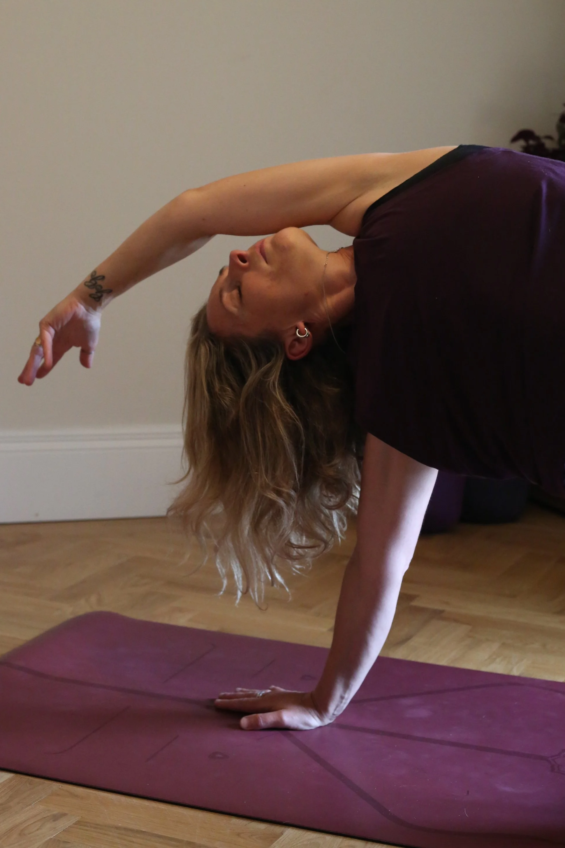 A woman performing a yoga pose in a room with wooden flooring, on a purple yoga mat, with her left arm supporting her body as she stretches her right arm over her head. She has long, wavy hair and tattoos on her left wrist.