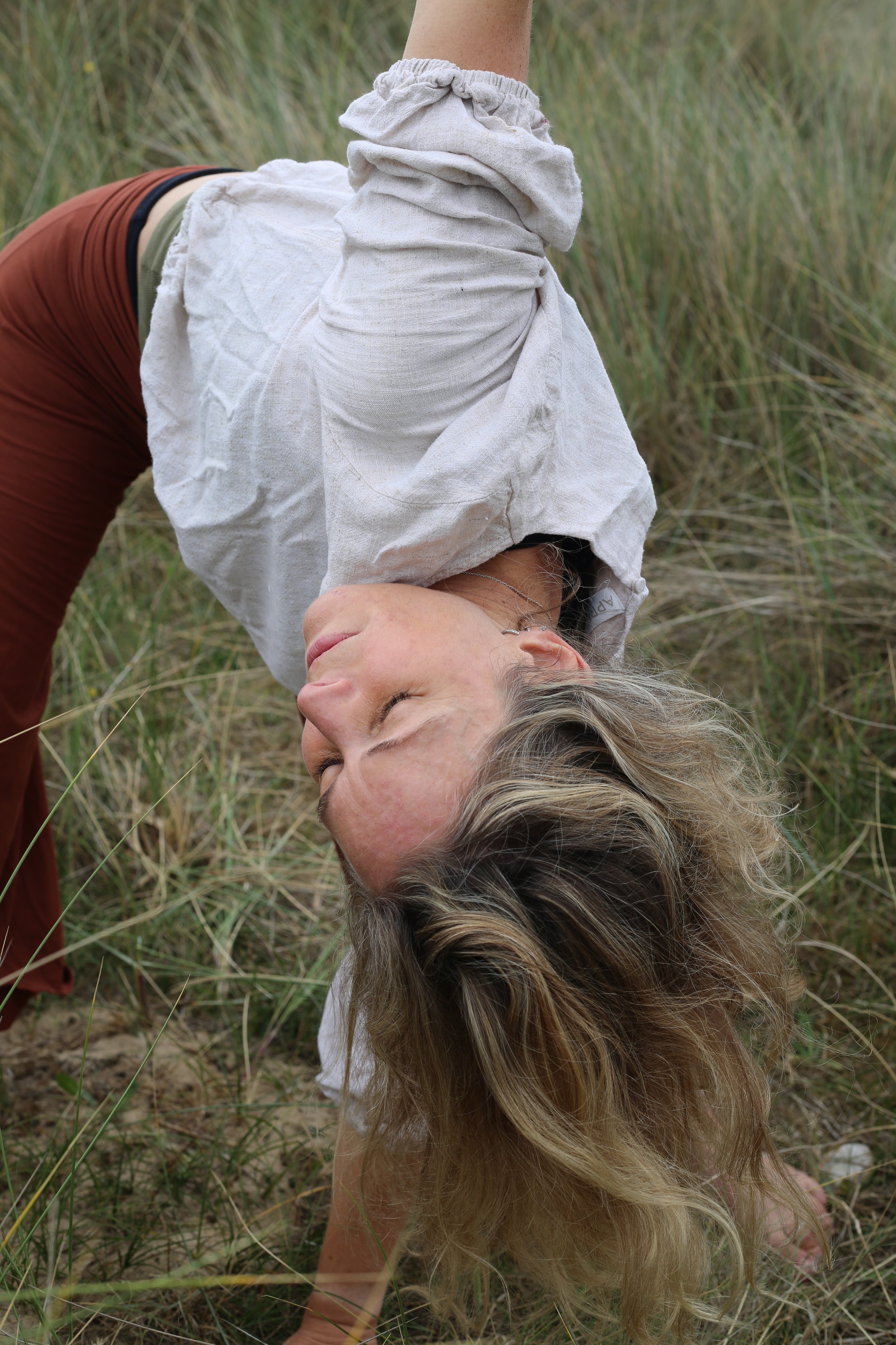 A woman with long blonde hair lying on the grass in a field of tall grass, wearing a white long-sleeve shirt and reddish-brown pants.