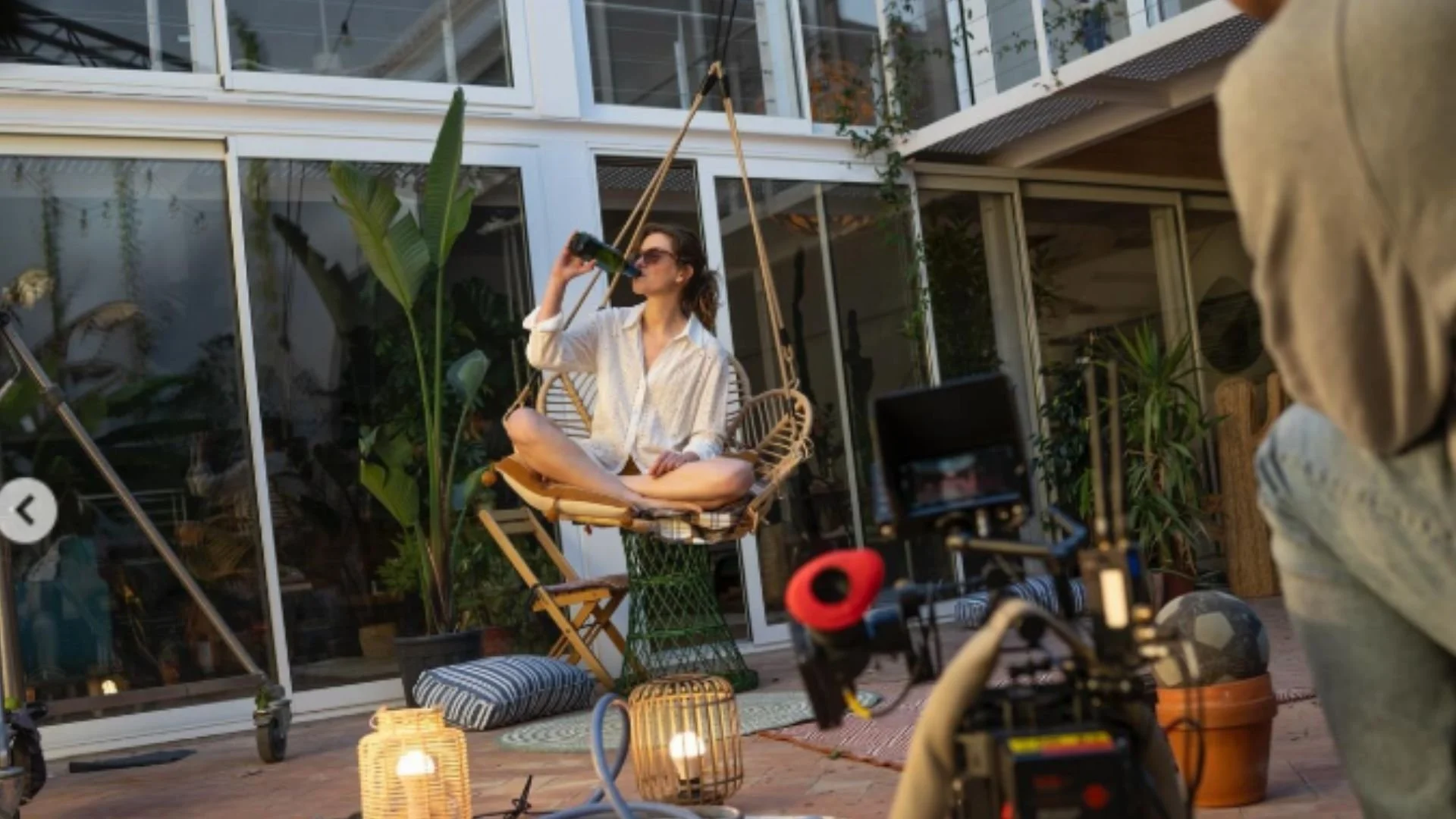 Woman sitting cross-legged on a swing chair, drinking from a wine bottle, during filming setup outside a modern house with large glass windows and plants.
