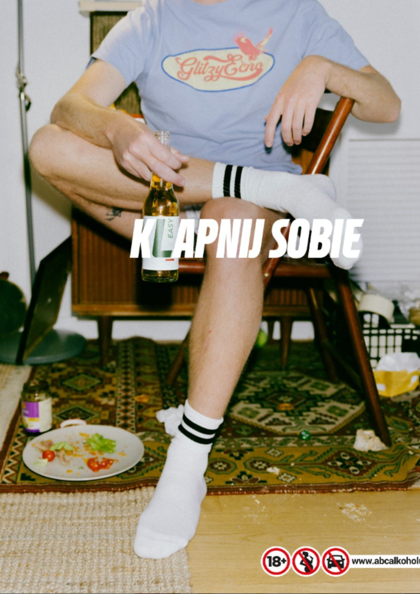 A person sitting on a wooden chair, holding a beer bottle, wearing a blue t-shirt with a 'GlizyGina' logo, white shorts, and white socks with black stripes. There are beer bottles, a plate with food, and trash on the floor, with a patterned rug. Text in the image says 'Klapnij Sobie' and symbols indicating age restriction, no minors, and no drinking and driving.