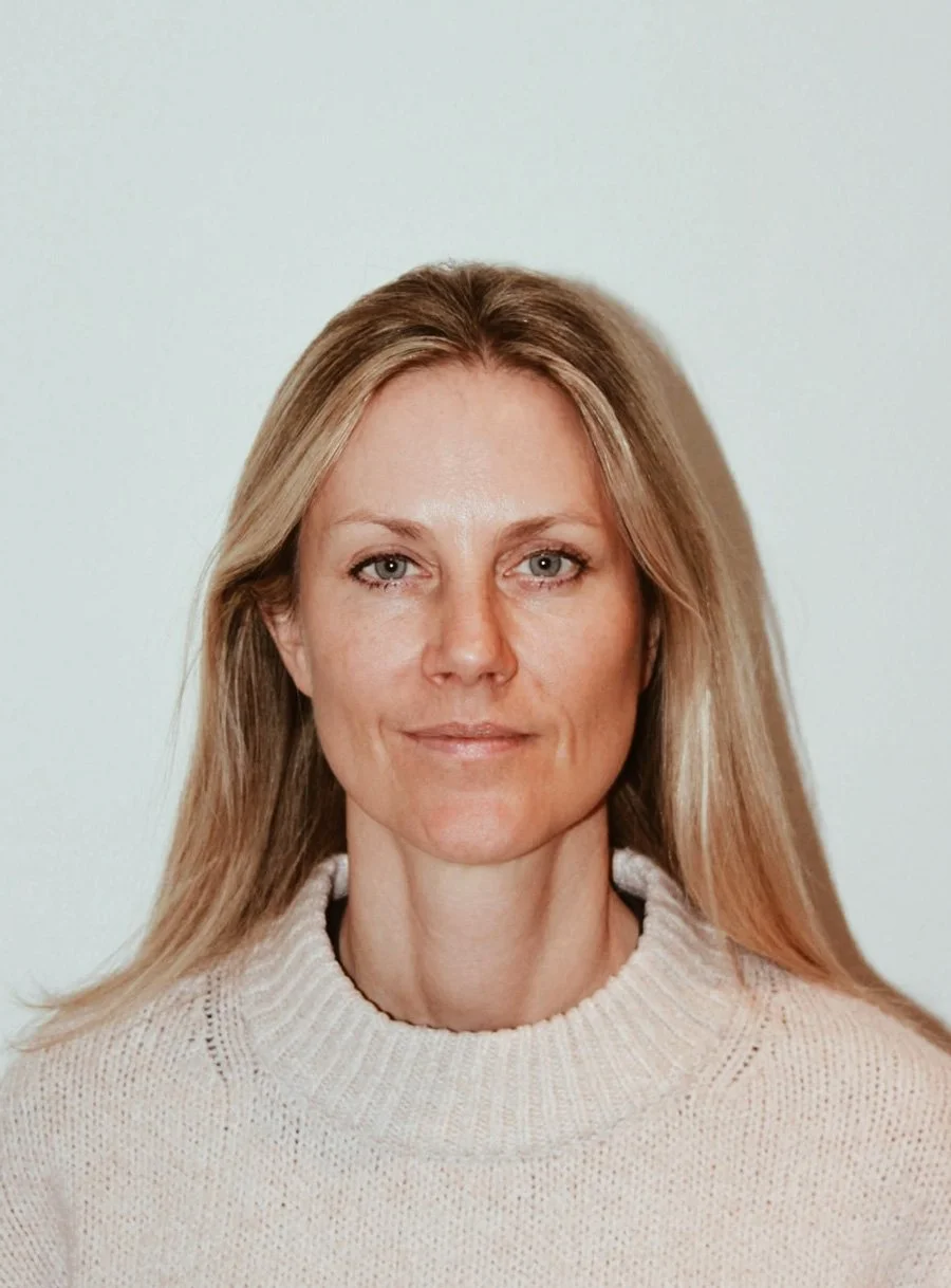 Porträtt av en kvinna med långt blont hår, klädd i en ljus beige stickad tröja, står mot en neutral bakgrund.