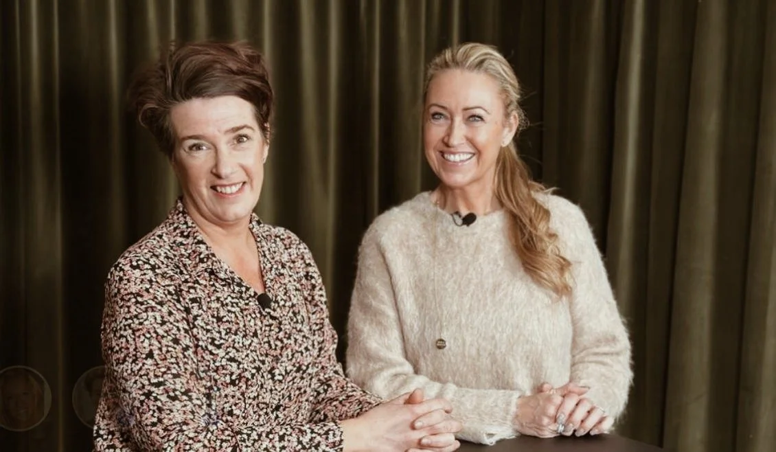 Två kvinnor som står och ler, den ena i en blommig blus och den andra i en ljus stickad tröja, framför bakgrund av gröna gardiner.