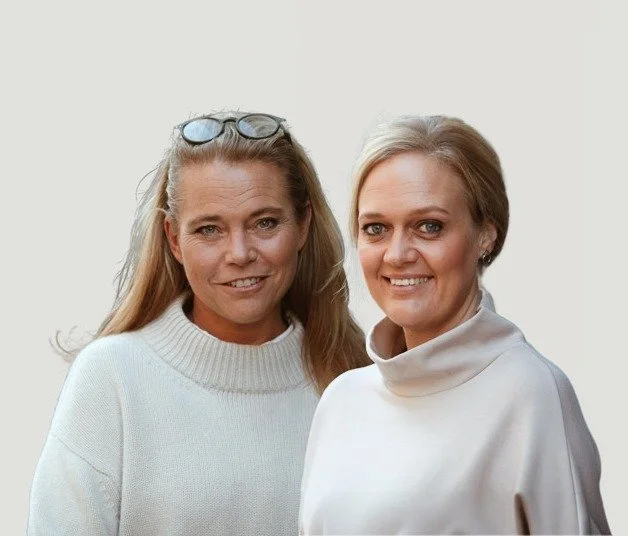 Två kvinnor står sida vid sida, båda ler och har ljus hår, den ena har glasögon på huvudet och den andra är klädd i en vit tröja.